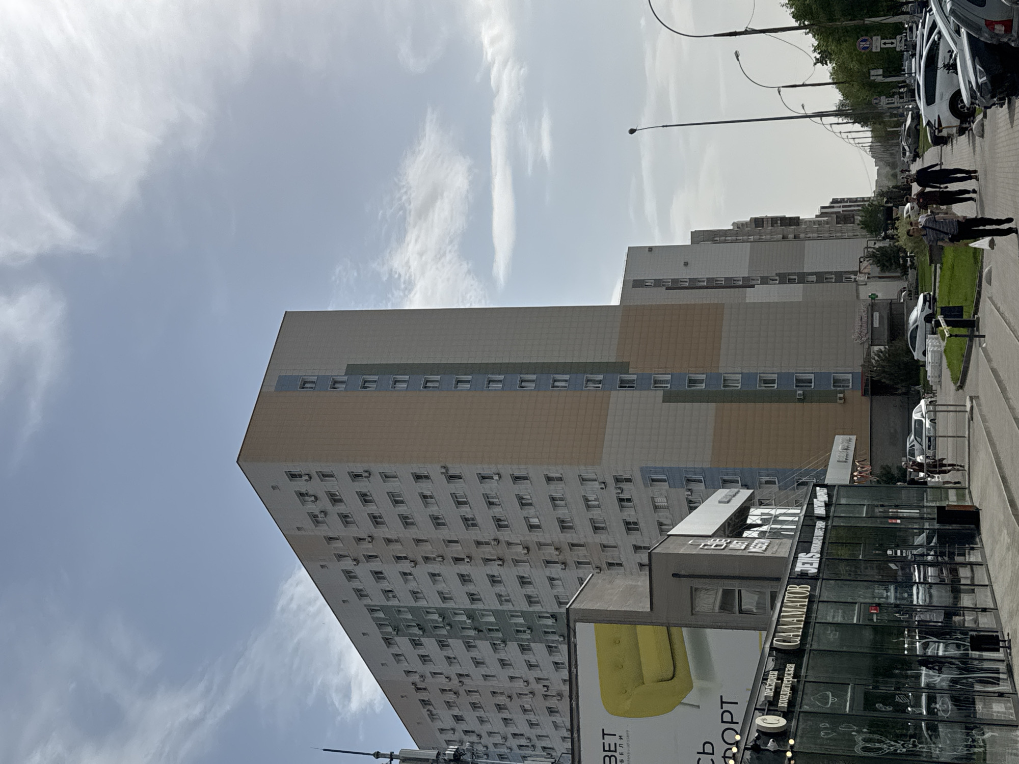 The image shows a tall, multi-story building in the city of Unknown, Unknown, viewed from a low angle. The building has a rectangular shape with numerous windows arranged in a grid pattern on its facade. The color scheme of the building includes gray, tan, and blue sections. In front of the building is a modern glass-walled structure with signage in Cyrillic script. The structure appears to be a commercial establishment. A billboard featuring a yellow design is positioned near the glass structure.
On the right side of the image, there is a sidewalk with people walking. Cars are parked along the street. The sky is visible in the background and is partially covered with clouds. Street lights are present along the street.