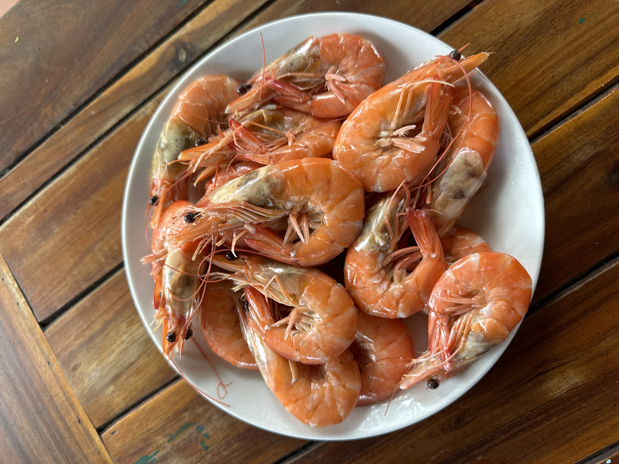 A top-down shot showcases a generous serving of cooked shrimp arranged on a white plate, set against a rustic wooden table. The plate is filled with numerous whole shrimp, their shells displaying a vibrant reddish-orange hue, indicating they have been boiled or steamed. Many of the shrimp retain their heads, with distinct black eyes and delicate, translucent antennae still attached. Their bodies are curled, a characteristic of cooked crustaceans.
A small amount of liquid, possibly from the cooking process, is visible at the bottom of the plate, along with a few dark specks that appear to be peppercorns, suggesting light seasoning. The white ceramic plate provides a clean contrast to the warm tones of the seafood.
The background consists of a wooden table made of planks, exhibiting natural grain and varying shades of brown. The planks appear worn, with visible seams and slight imperfections. Faint traces of what might be green and blue paint or residue are discernible on some parts of the table surface, suggesting a casual or well-used environment. The scene depicts a ready-to-eat meal, likely fresh seafood, fitting for a coastal location such as Mũi Né, Vietnam. There are no people or discernible text in the image.