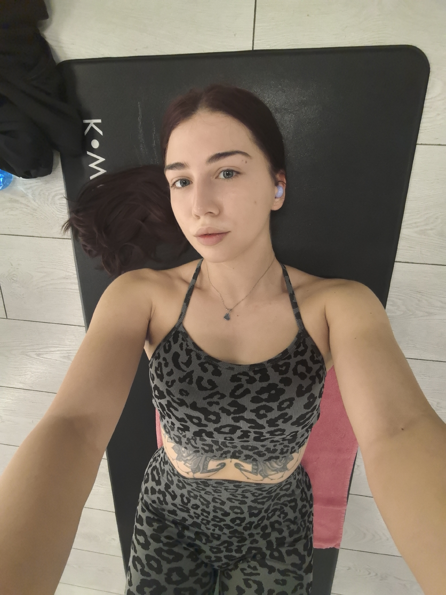 A young woman lies on a black exercise mat in an indoor setting, likely a home gym. She is wearing a grey and black leopard-print sports bra and matching leggings. She has brown hair, fair skin, and appears to be wearing light purple earbuds. A necklace with a small pendant is visible around her neck. A tattoo is visible on her abdomen. Part of a dark-colored jacket or garment can be seen in the upper left corner of the frame. A pink towel is visible beneath her midsection. The floor is light-colored with a wood-grain pattern. The letters "K•M" are printed on the exercise mat near the upper left.
The location in the city of Stara Zagora, Bulgaria, cannot be confirmed from the information available.