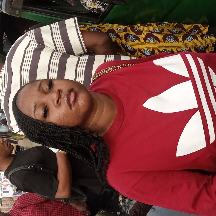 A young woman with long braids is wearing a red Adidas shirt with a gold chain strap visible on her shoulder. She looks directly at the camera with a slight pout. The background is a busy street scene with pedestrians, and a vehicle interior with striped and patterned upholstery. The angle suggests the photo was taken while she was lying down or in a reclining position.