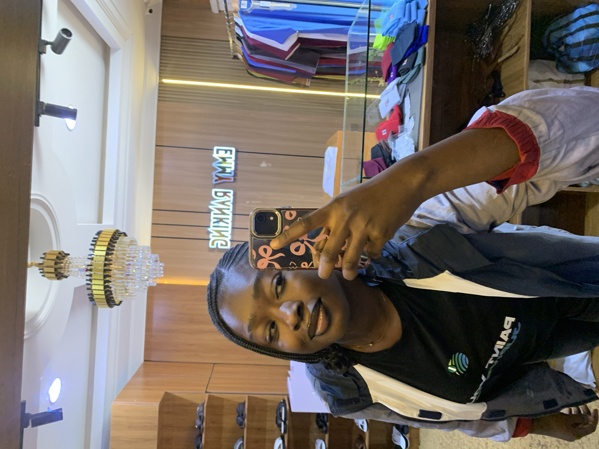 A young Black woman with braided hair is taking a mirror selfie in what appears to be a clothing store. She is wearing a black t-shirt with a colorful logo and a grey and white jacket with red trim on the sleeves. She is holding up a smartphone with a pink and black case. In the background, there are shelves stocked with shoes and clothing, as well as a neon sign that reads "EMMY RANKING." The store is well-lit with a chandelier and track lighting. The image was taken in Gura Top, Nigeria.