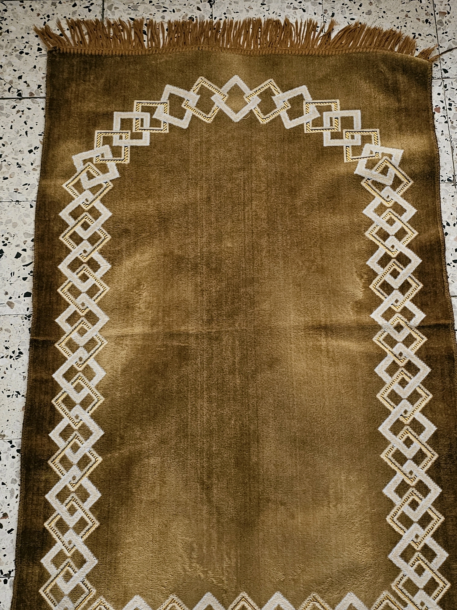 A brown prayer rug with a geometric white and gold border is laid out on a speckled floor. The rug has a fringe along the top edge. The geometric pattern consists of interlocking squares. The rug is positioned horizontally in the frame, occupying the majority of the image. The background appears to be a terrazzo-style floor with black and white chips. The image is a top-down view. This image does not provide any information about the city of Batna, Algeria.