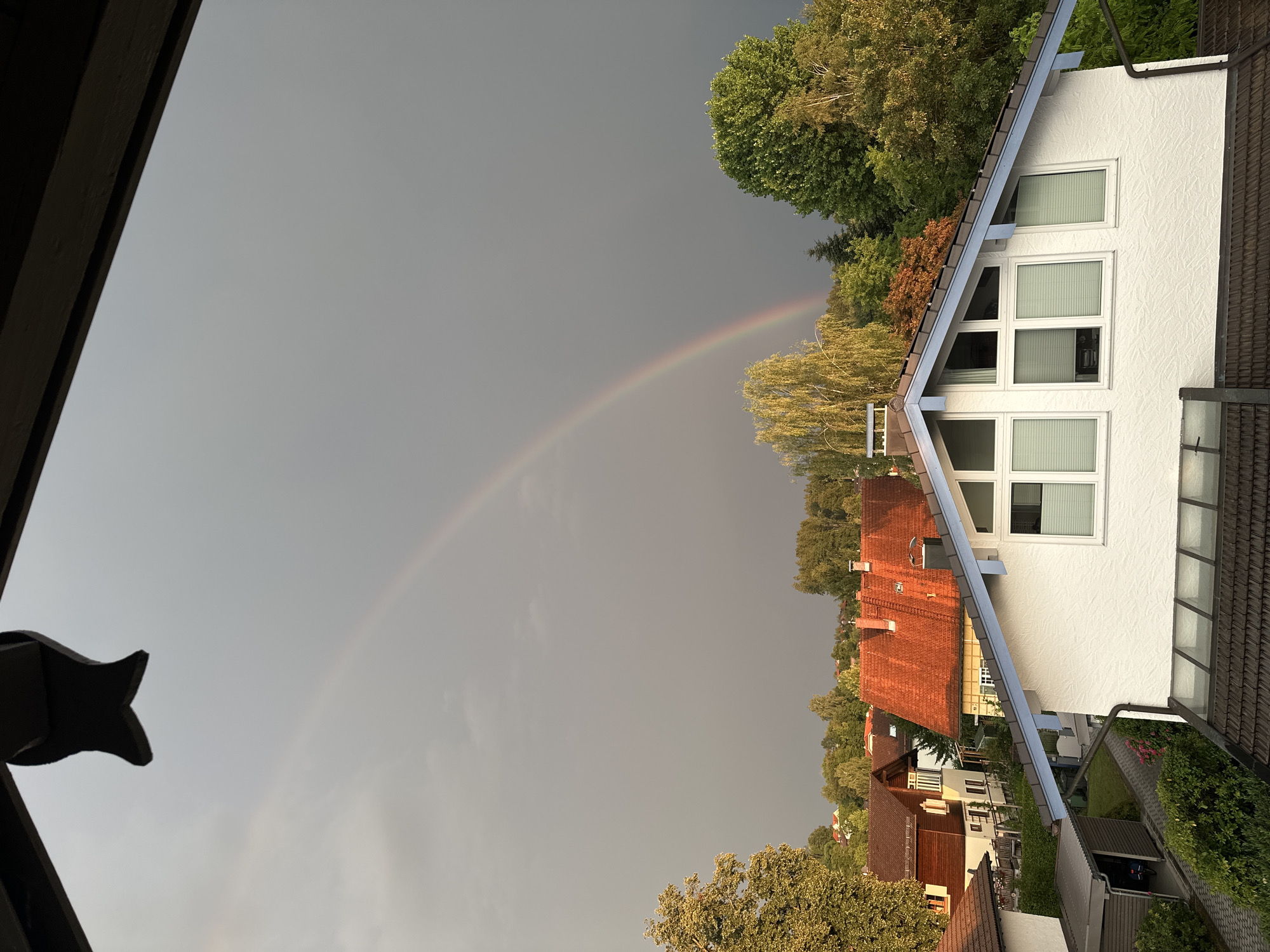 The image depicts a suburban scene in Bad Endorf, Germany, under a dramatic, overcast sky, with a faint rainbow arcing across the upper portion of the frame. The foreground is dominated by the roofline of a modern white house with a prominent blue-trimmed gable and multiple windows fitted with white blinds. Below this, a darker tiled roof is visible, along with a portion of a grey shed containing a bicycle.
Behind the foreground house, a collection of other residential buildings with traditional red-tiled roofs are nestled amongst lush green trees. These houses appear to be varied in architectural style, with some featuring wooden elements. The trees are dense and create a sense of a well-established residential area with significant greenery. The overall lighting suggests late afternoon or early evening, with the overcast conditions casting a diffused light over the landscape, punctuated by the subtle hues of the rainbow. There are no people or animals visible in the scene.