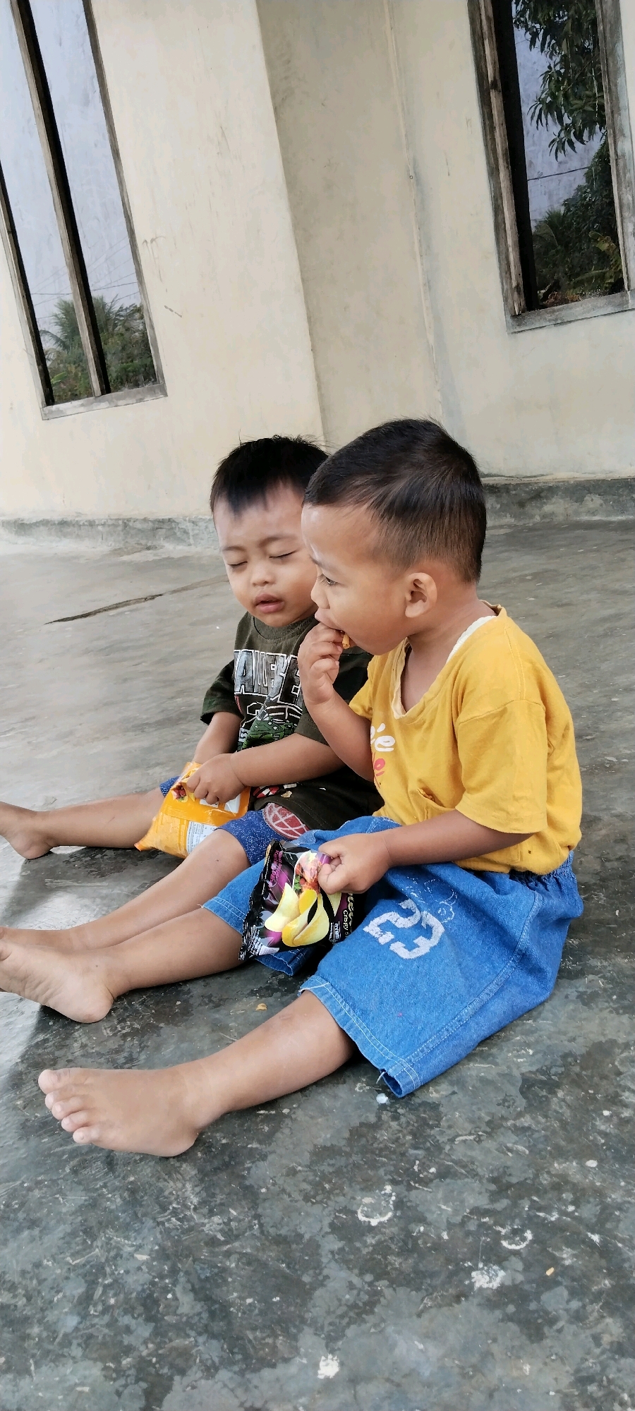 Two young boys are sitting on a grey, concrete floor. The boy on the left, wearing a dark green t-shirt with a white graphic, has his eyes closed and is holding a yellow snack bag in his left hand. The boy on the right, wearing a yellow t-shirt and blue denim shorts with a white number "23" printed on the right leg, is looking to his left and eating a snack. He is holding a snack bag with images of sliced fruit. Both boys are barefoot. The background shows a light-colored, stucco wall with two windows. The window on the left has reflections of palm trees and what appears to be a cloudy sky. The window on the right shows foliage and a grey structure. The scene is outdoors, likely in a residential or communal area. The image was taken in Tanjung Ratu Ilir, Indonesia.