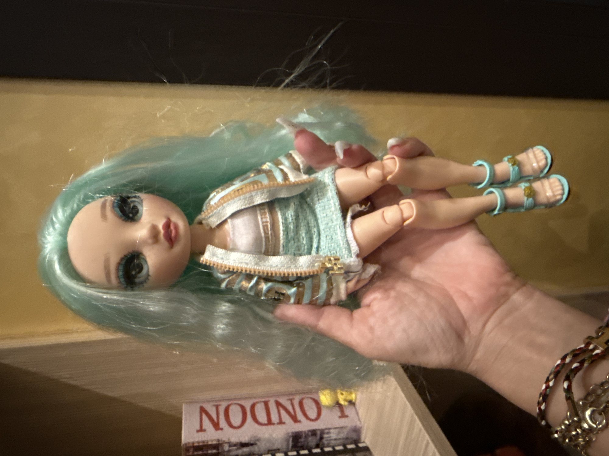 The image shows a close-up of a doll with light blue hair and striking blue eyes, held in a person's hand. The doll is wearing a textured light blue and gold striped jacket over a white top and a matching light blue skirt. Its feet are adorned with light blue sandals. The doll's fingers have long, white nail polish. The person's hand is supporting the doll, and their wrist is decorated with several bracelets, including a braided one with red, white, and blue threads, and a silver chain bracelet.
In the background, on a shelf-like surface, there is a small item with the word "LONDON" printed in red, stylized letters, with an image of a train beneath it. The lighting suggests an indoor setting, likely with artificial light, and the background is blurred, focusing attention on the doll. The overall impression is a casual, personal photograph, possibly a snapshot of a toy or collection. The presence of the "LONDON" item provides a subtle hint of travel or interest in the city.