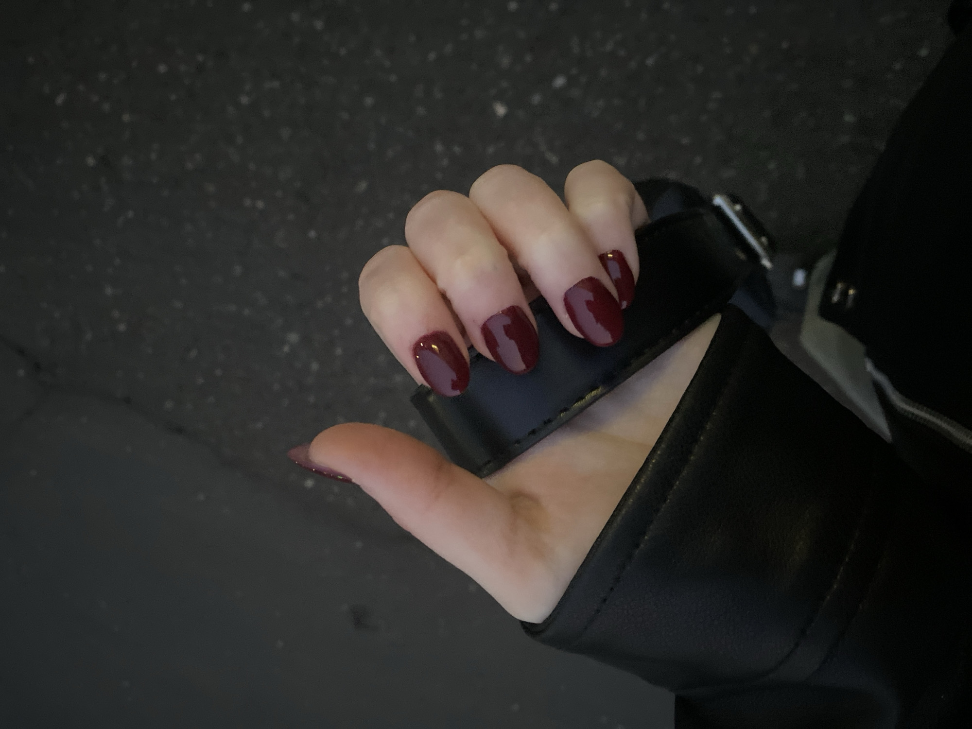 A hand with short, dark red polished nails is gripping a black leather bag strap. The person is wearing a black leather jacket. The background shows a dark grey asphalt surface. The image does not provide information about the location, nor if it's in the city of Izhevsk, Russia.