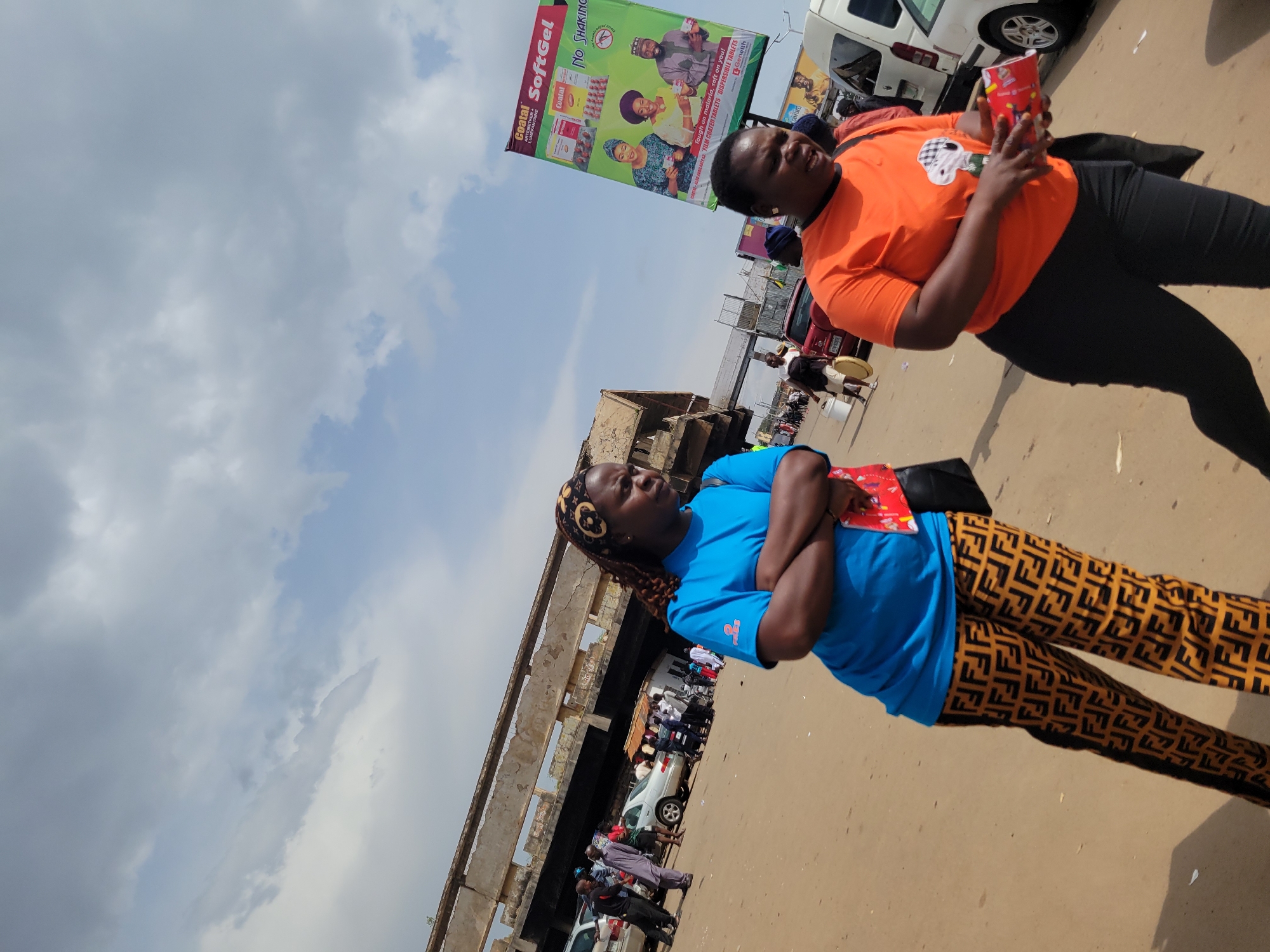 An outdoor scene in the city of Jos, Nigeria features a dusty, unpaved ground with several people and vehicles. In the foreground, two women are prominent. The woman on the left is wearing a bright blue t-shirt and patterned pants, with her arms crossed. She has dark hair pulled back in braids and a patterned headband. The woman on the right is wearing an orange t-shirt and black pants, holding a red box in her hands. She has short, dark hair and is looking to her right, with her mouth slightly open.
In the background, there is a large, dilapidated concrete structure, possibly an overpass or an unfinished building, with exposed rebar. A billboard advertising "Coatal SoftGel" with images of people is visible on the right side of the image. Several cars, including a red car and white van, are parked or driving in the area. Other people are scattered throughout the scene, some walking and some standing in groups. The sky is partly cloudy with a mix of grey and blue. The overall impression is of a busy, somewhat chaotic urban environment.