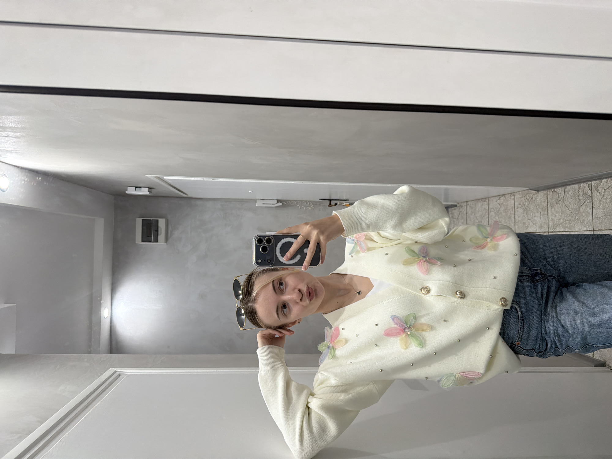 A young woman is taking a selfie in a mirror, likely in a bathroom or similar space. She is wearing a white cardigan adorned with colorful floral embellishments and blue jeans. Sunglasses are perched on her head. The background reveals a simple interior with white walls and what appears to be light gray tiling. Although it is impossible to determine the exact location in Plovdiv, Bulgaria, from the image alone, one can assume that she is in a private or semi-private space within the city. The primary action is her taking a selfie, using her phone, which is positioned in front of her face. There is no visible interaction with any other individuals or significant elements within the environment.