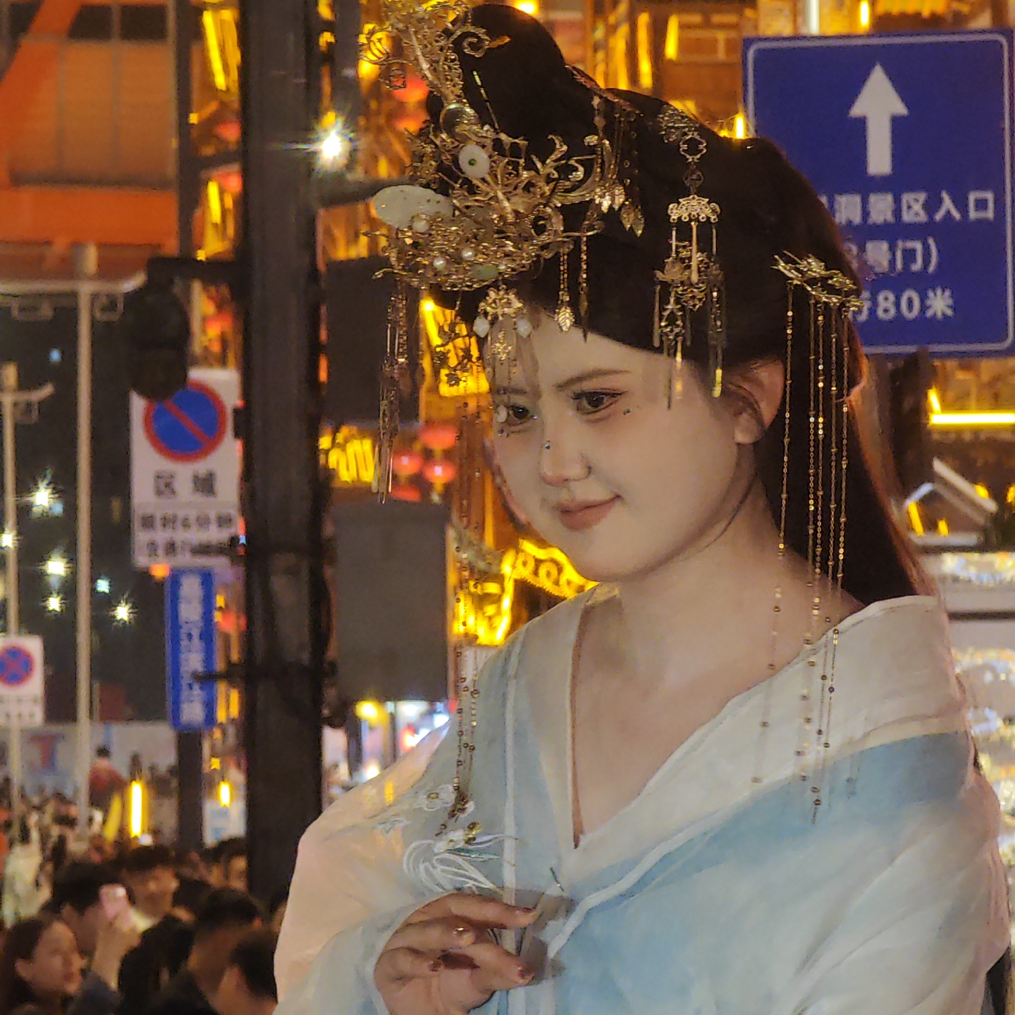 A woman in traditional Chinese attire stands in a busy urban environment. She has black hair adorned with ornate gold jewelry and is wearing a light blue and white robe. In the background, there are street signs in Chinese, traffic lights, and numerous bright lights that suggest an evening setting. A crowd of people is visible behind her, indicating a public gathering. The general location is the Yuzhong District in China, as suggested by the street signs.
