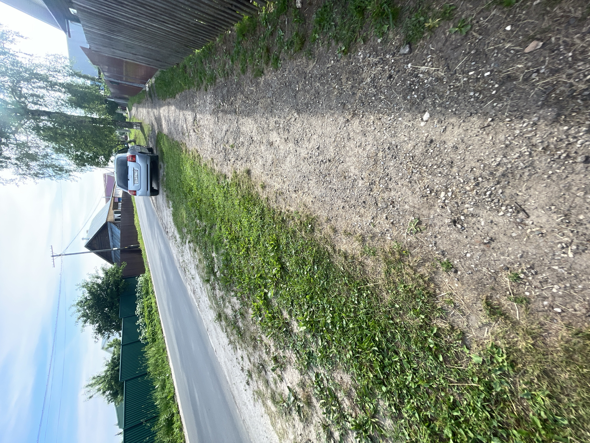 The image shows a rural road in Krasnye Baki, Russia, with a dirt path running alongside it. The road is paved with asphalt and bordered by a green metal fence on the left. The path on the right is made of gravel and dirt, with patches of grass growing along its edges. Several houses and trees are visible in the background, and a silver car is parked on the side of the road near the path. The sky is blue with scattered clouds, and the sun is shining, casting shadows on the ground. The overall scene depicts a quiet, everyday street in a Russian village.