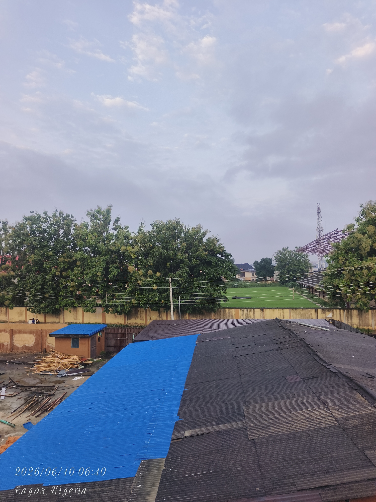 The image depicts an aerial view of a construction site in Shangisha, Nigeria, on June 10, 2026, at 06:40. In the foreground, two sections of rooftops are visible: one with a bright blue corrugated metal roof and another with a darker, weathered corrugated metal roof. Piles of lumber and construction materials are scattered on the ground near a small, orange-brown structure with a blue roof. In the midground, a tall concrete wall separates the construction area from a green field, possibly a sports field, with stadium seating and a transmission tower in the distance. Lush green trees line the perimeter of the field. The sky is overcast with scattered clouds, suggesting early morning or a cloudy day. Two indistinct figures are visible on top of the concrete wall in the distance.