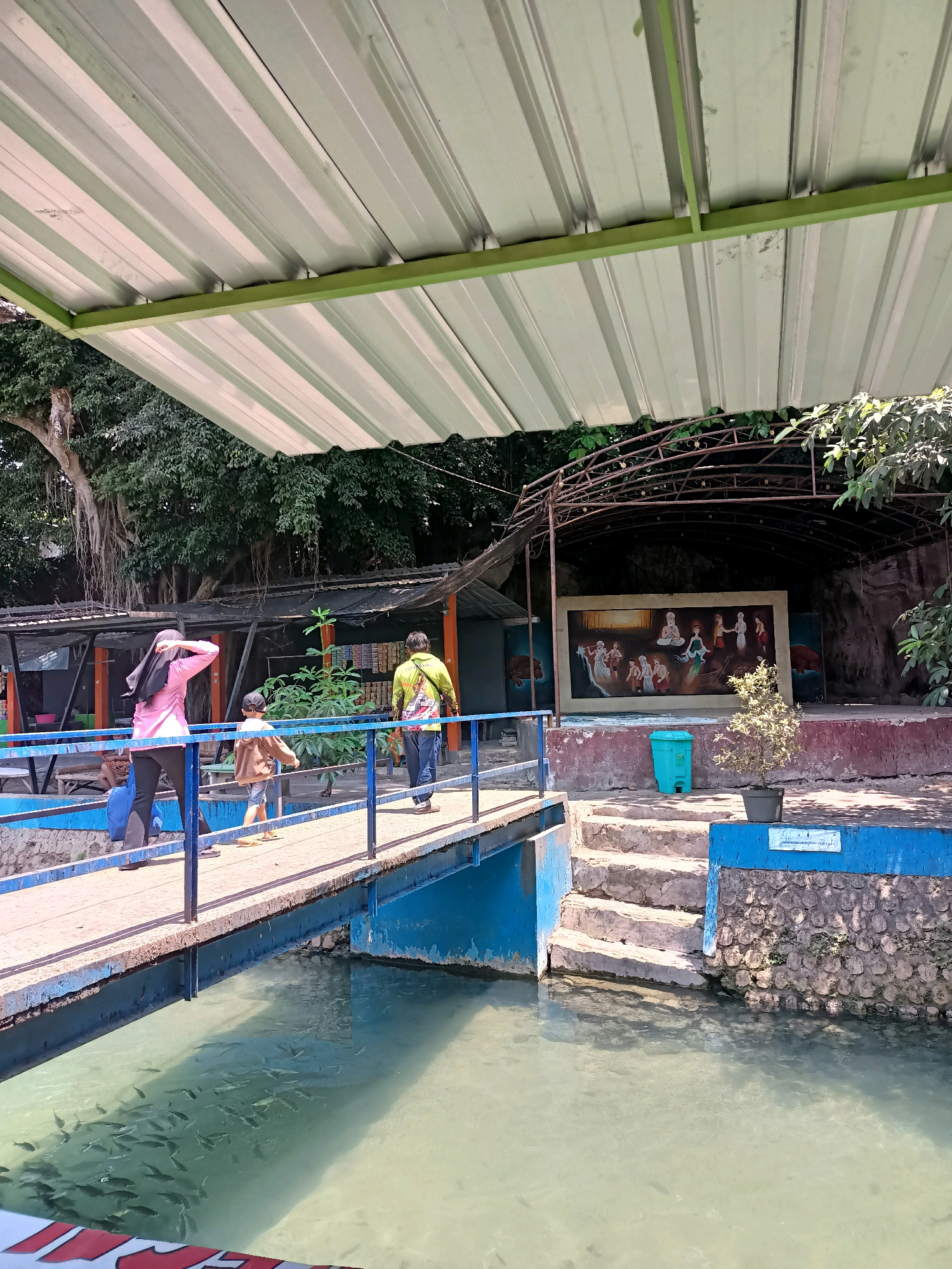 The image captures a scene at a water attraction, likely a tourist spot, in Rengel, Indonesia. A bright blue and white metal roof structure with supporting green beams covers a significant portion of the upper frame. Below this, a wooden walkway with blue railings spans over a body of water teeming with fish. Three people are walking on the bridge: a woman in a pink hijab and dark pants, a child in a brown jacket and blue shorts, and a man in a yellow-green shirt and blue jeans.
To the right of the walkway, stone steps lead up to a stage area. A large painting depicting a scene with several figures is displayed on the stage. A turquoise trash can and a potted plant are positioned near the steps. The background reveals dense green trees, and some stalls or small buildings are visible behind the walkway. The water appears murky and reflects the overhead structure. The overall environment suggests an outdoor recreational area with a focus on water and potentially cultural or artistic elements on the stage.
