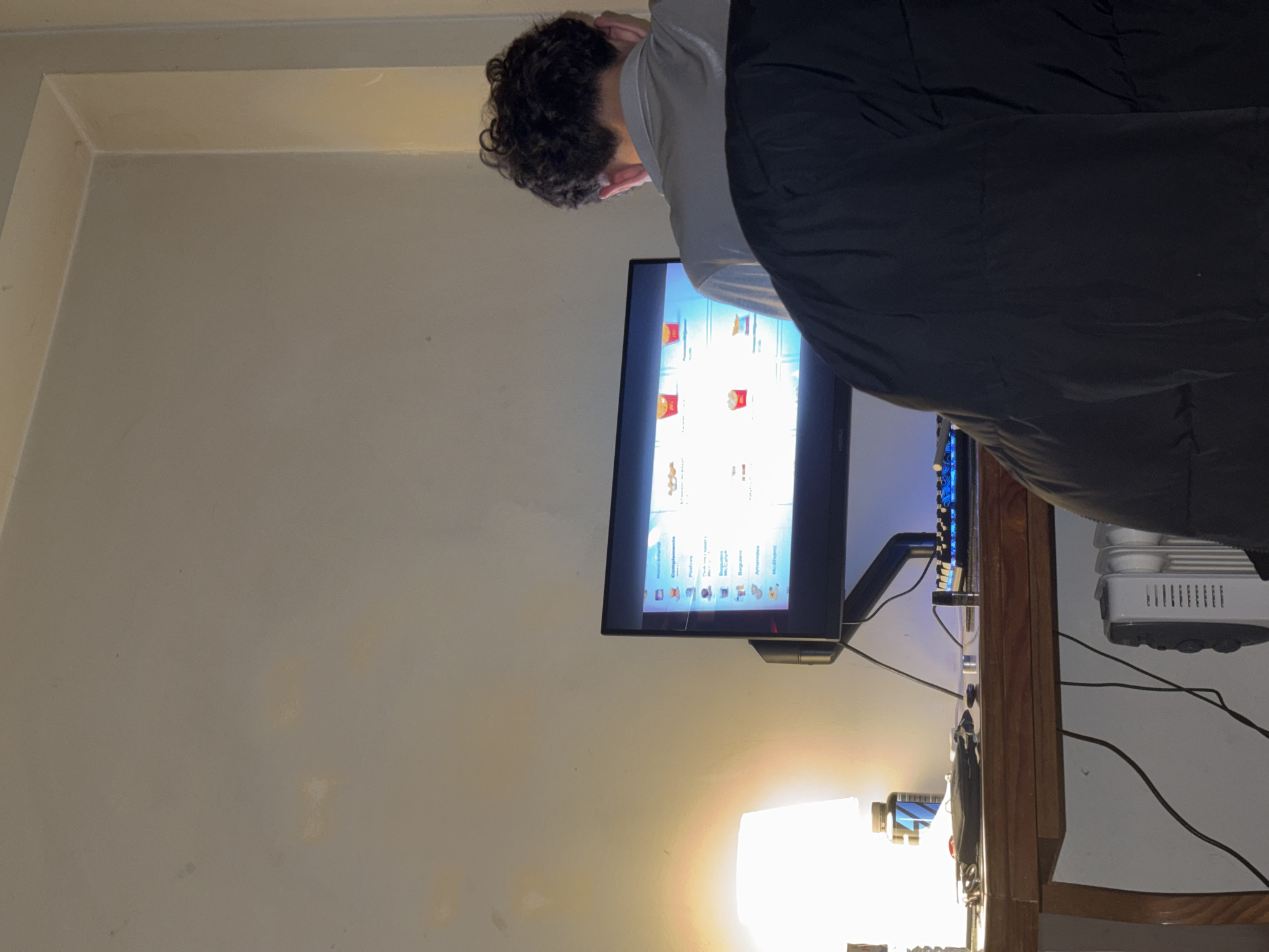 An indoor scene captures a person engaging with a computer monitor, likely in an evening or nighttime setting. The image is oriented vertically but depicts a person with dark, curly hair, seen from behind and the side, leaning slightly forward as they face a brightly lit screen. They are wearing a light-colored top and dark clothing.
The setting appears to be a room, possibly a bedroom or home office, with plain, light-colored walls. A wooden desk or table is positioned beneath the monitor. On the desk, a lamp emits a warm, bright glow, providing a significant light source in the otherwise dimly lit room. To the right of the monitor, a white electronic device with visible vents and controls, resembling a humidifier or small air purifier, sits on the desk. Numerous cables are strewn across the desk surface, connecting the various electronic items.
The computer monitor displays a graphical user interface, which appears to be a digital menu or ordering system, possibly from a fast-food establishment. Several food icons are visible, including what look like french fries in red containers and various beverages. Prominently displayed text on the screen, in Portuguese, includes categories such as "Menu Infantil" (Children's Menu), "Complementos" (Complements), "Pratos" (Dishes), "Bebidas" (Drinks), "Acompanhamentos" (Accompaniments), and "Sobremesas" (Desserts). The overall impression is one of someone browsing or selecting items from a digital menu.
