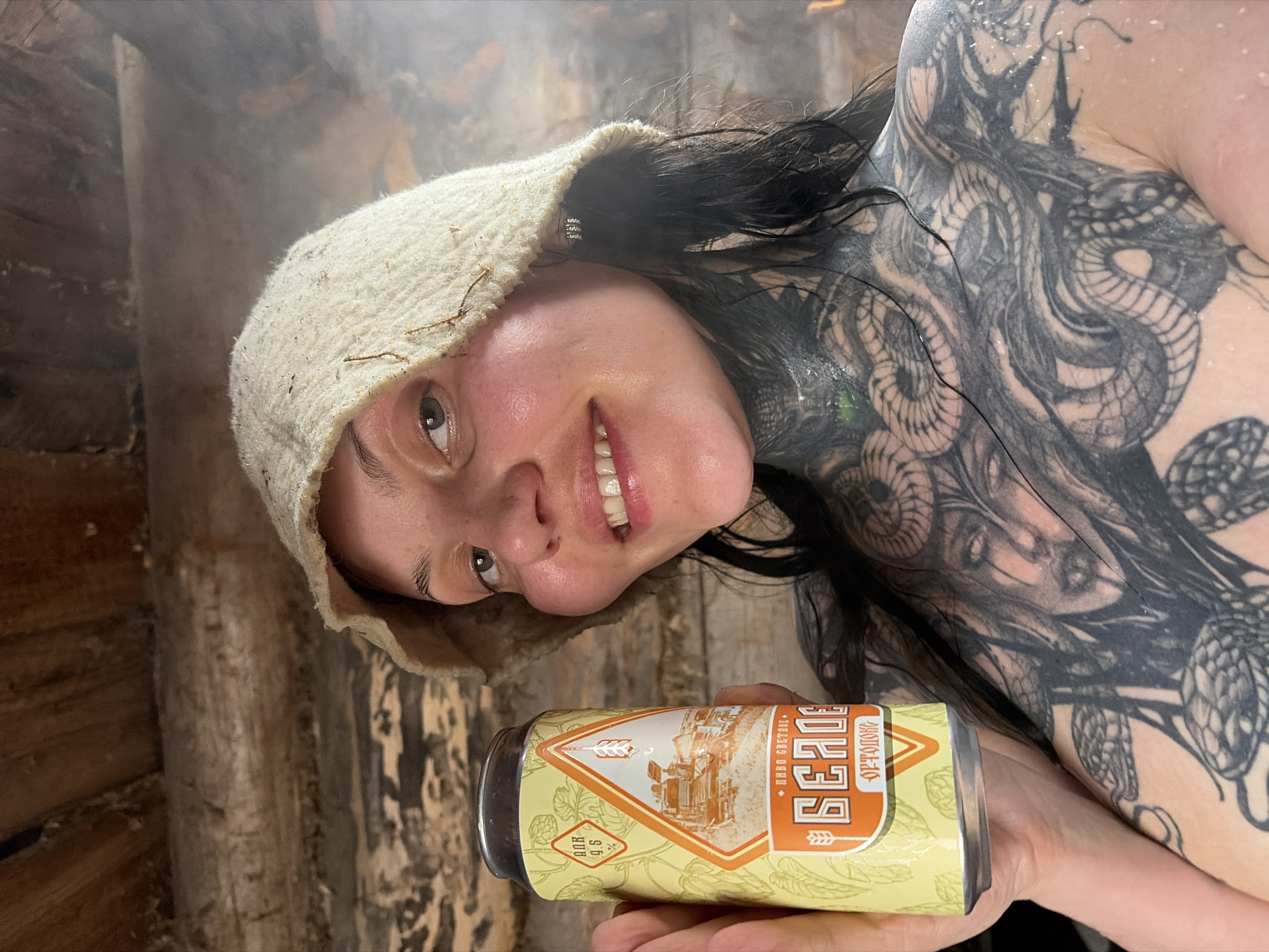 A person, presumably female, is depicted holding a yellow can of "БЕЛОЕ ЗОЛОТО" beer. The individual is wearing a white hat and has dark hair, fair skin, and tattoos covering their shoulder and chest. Water droplets are visible on their skin. The background suggests a wooden structure, possibly a sauna or a cabin, and the scene is foggy. The location of the scene is not explicitly mentioned, but the image contains reference to Буренка, Russia.