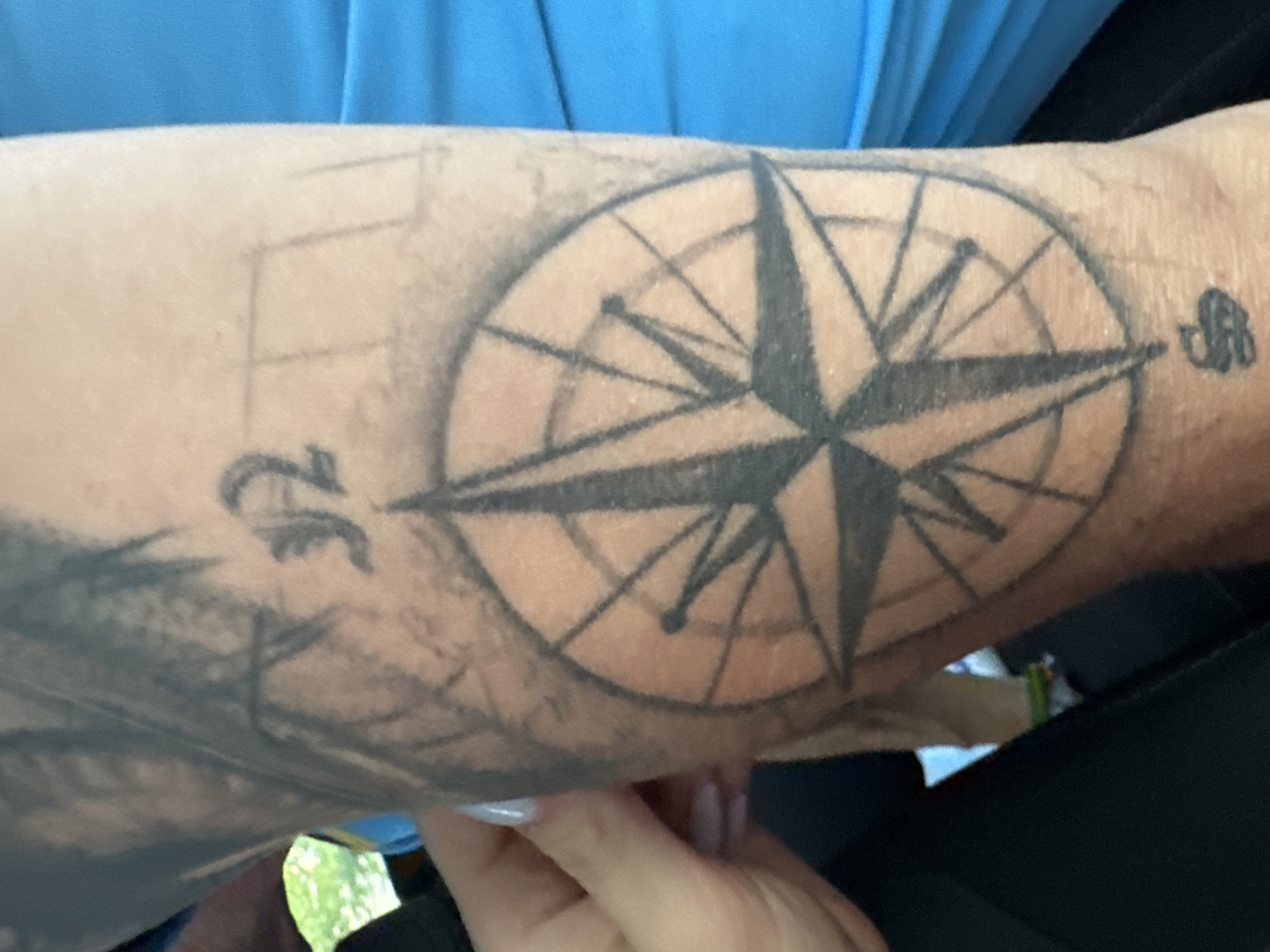 The image shows a close-up of a person's arm with a tattoo of a compass rose. The tattoo is black and gray, and it appears to be a recent addition or an unfinished piece, as there are faint sketch lines visible around it. Above the compass rose, there is another smaller tattoo that looks like a stylized letter or symbol. The person's skin appears fair. To the left of the arm, a hand with manicured nails is visible, suggesting the person is wearing a blue shirt. The background is blurred, but it seems to be an outdoor setting with greenery, possibly a park or a street scene. The image does not provide any information about the city of Murcia, Spain, or any other specific location.