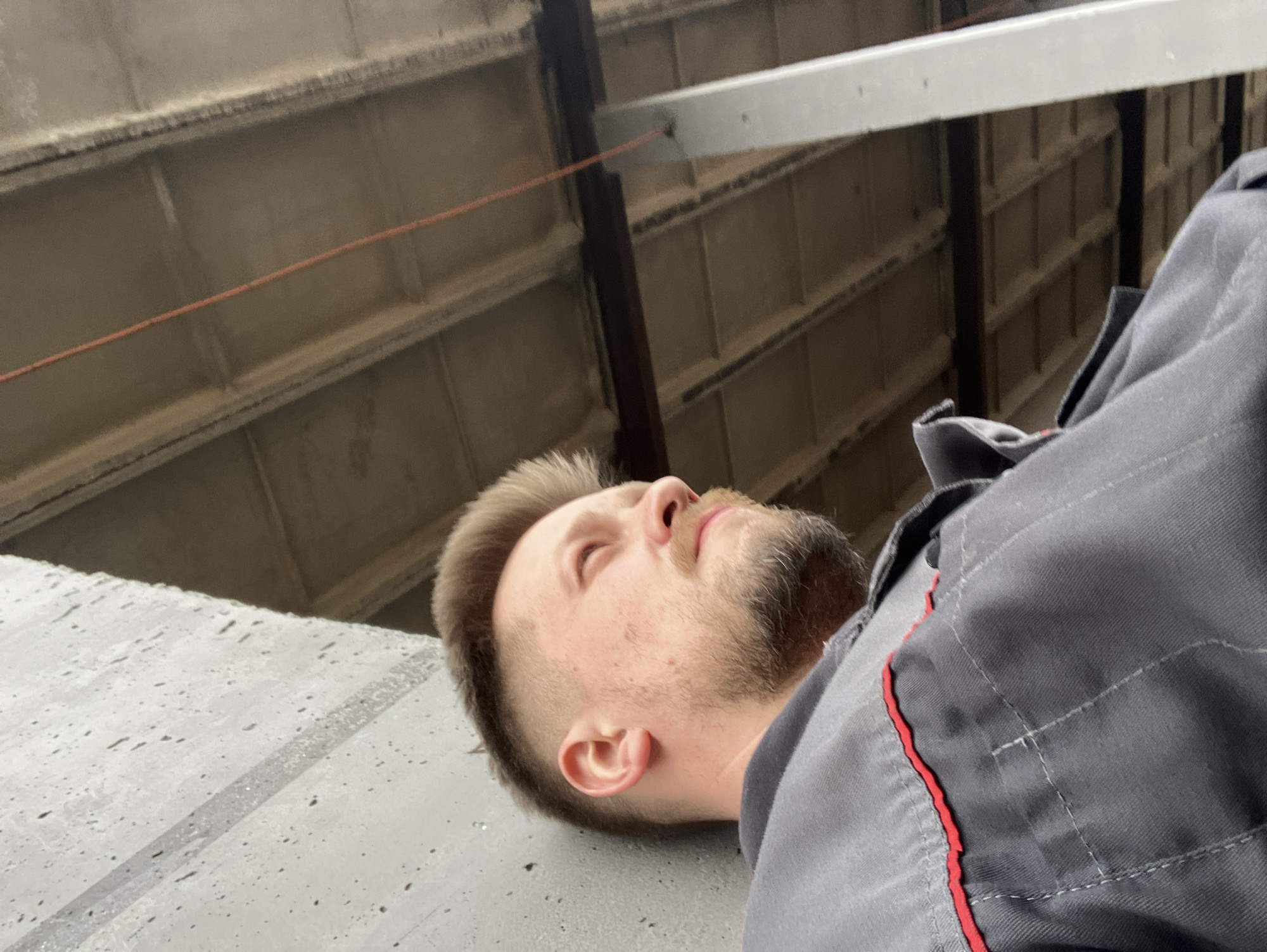 The image features a man with light brown hair and a beard, seen from the chest up, lying on his side and looking upwards with his eyes closed. He is wearing a dark gray collared shirt with red trim and white stitching, suggesting a work uniform. The surface he is resting on is light gray, textured, and speckled, possibly concrete or metal, with some faint streaks that could indicate moisture.
Above him, the background reveals an industrial-style ceiling structure composed of a grid of light-colored rectangular panels or beams, interspersed with darker vertical support beams. An orange electrical wire runs horizontally across the mid-background, and a prominent white rectangular bar or beam is visible towards the top.
The setting appears to be an indoor or covered industrial environment, such as a workshop, garage, or factory. The general illumination suggests it is daytime. The man's closed eyes and relaxed posture convey a sense of rest or contemplation. There is no visible text in the image. The location is Noviy, Russia.