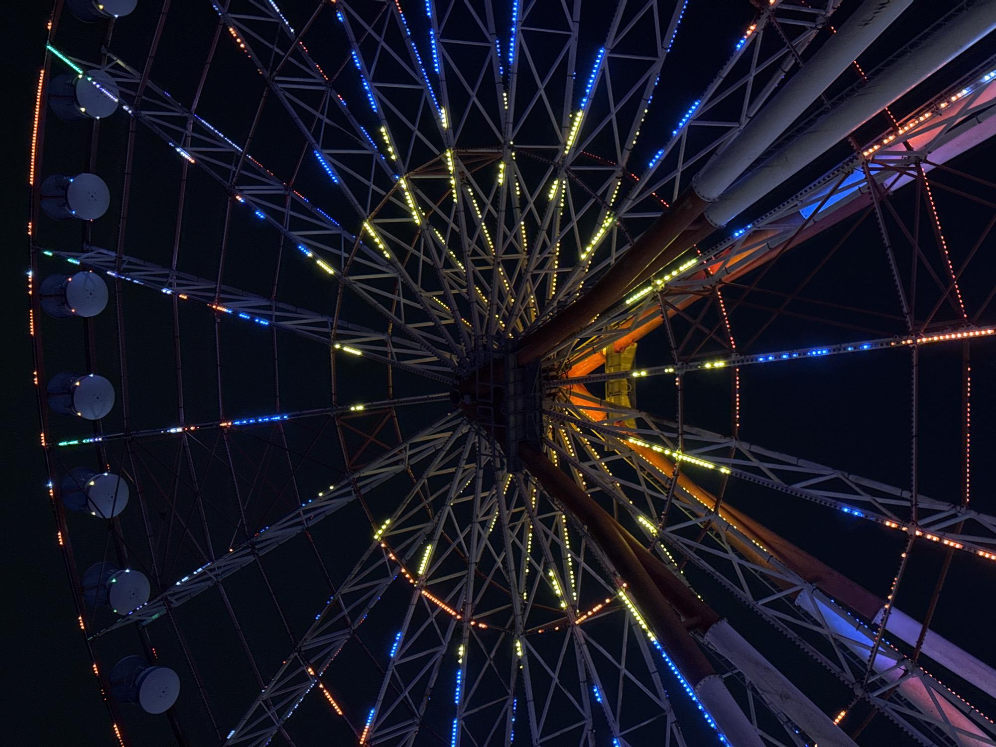 The image shows a view looking up at the Batumi Ferris Wheel in Batumi, Georgia at night. The Ferris wheel is illuminated with a variety of colorful lights, including blue, yellow, and orange. The structure of the wheel is visible, with its spokes and supporting beams. Several gondolas are attached to the rim of the wheel, also lit up with lights. The dark night sky serves as the background, emphasizing the bright lights of the amusement ride.
