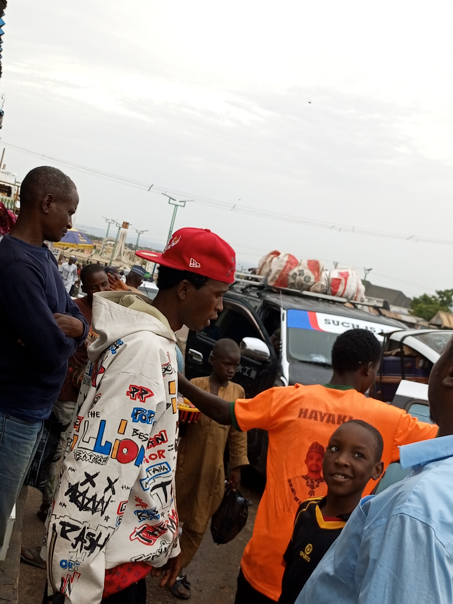 This image depicts a bustling scene, likely in the city of Gombe, Nigeria, possibly near a market or transportation hub. In the foreground, a young boy with a bright smile is the focal point, wearing a black and yellow sports shirt with the word "SPORTS" visible. He is standing next to an adult wearing a light blue shirt. To the left, a man in a dark blue long-sleeved shirt and jeans stands with his arms crossed, looking down. Another man, wearing a white hoodie adorned with colorful graffiti-style lettering, including words like "RASH" and "BATMAN," is the central figure in the middle ground. He wears a red baseball cap. Behind him, a dark SUV or van is visible, loaded with sacks on its roof, suggesting travel or transport. Several other individuals, including children and adults, are present in the background, some in traditional attire. The environment appears to be outdoors, with a cloudy sky overhead. The overall impression is one of everyday life and activity in an urban setting.