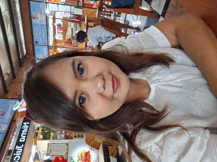 A woman with dark hair and fair skin is smiling at the camera. She's wearing a white textured top. The setting appears to be a casual restaurant or food court, as evidenced by the tables, chairs, and signage for "Nisnu Juice" in the background. A person in a gray shirt is visible in the distance behind her. The image is slightly rotated counterclockwise.