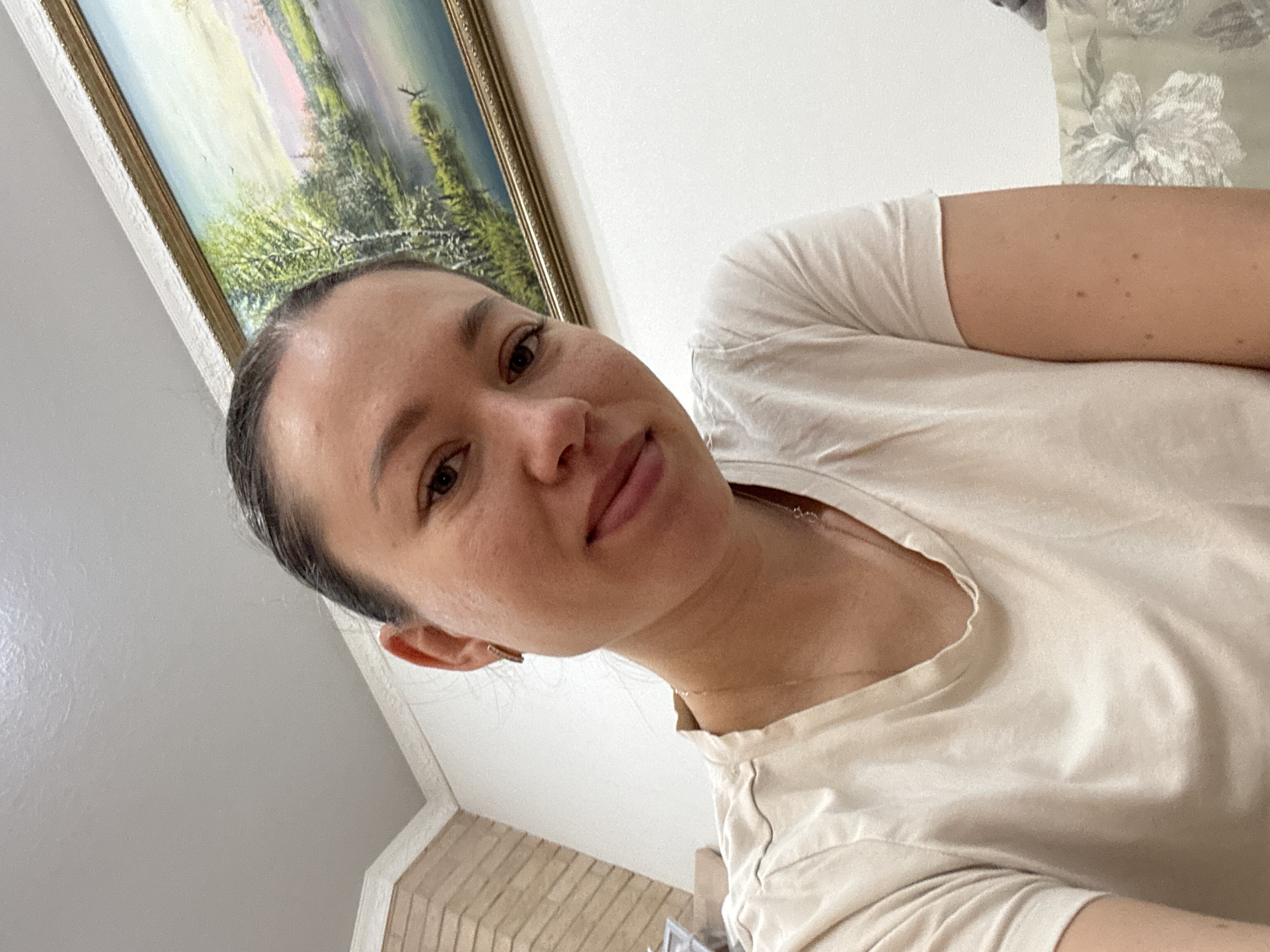 A woman with dark hair pulled back is pictured in a close-up shot. She has fair skin, brown eyes, and is wearing a light-colored t-shirt. A gold necklace is visible around her neck. In the background, a framed landscape painting hangs on a white wall. To the right, a floral-patterned pillow or cushion can be seen. The bottom of the image shows a portion of a brick wall or surface, suggesting the scene is indoors. The photograph appears to have been taken at an angle, possibly as a selfie. It's impossible to confirm the location as Ферма, Russia, as the image lacks specific details that would verify the setting.