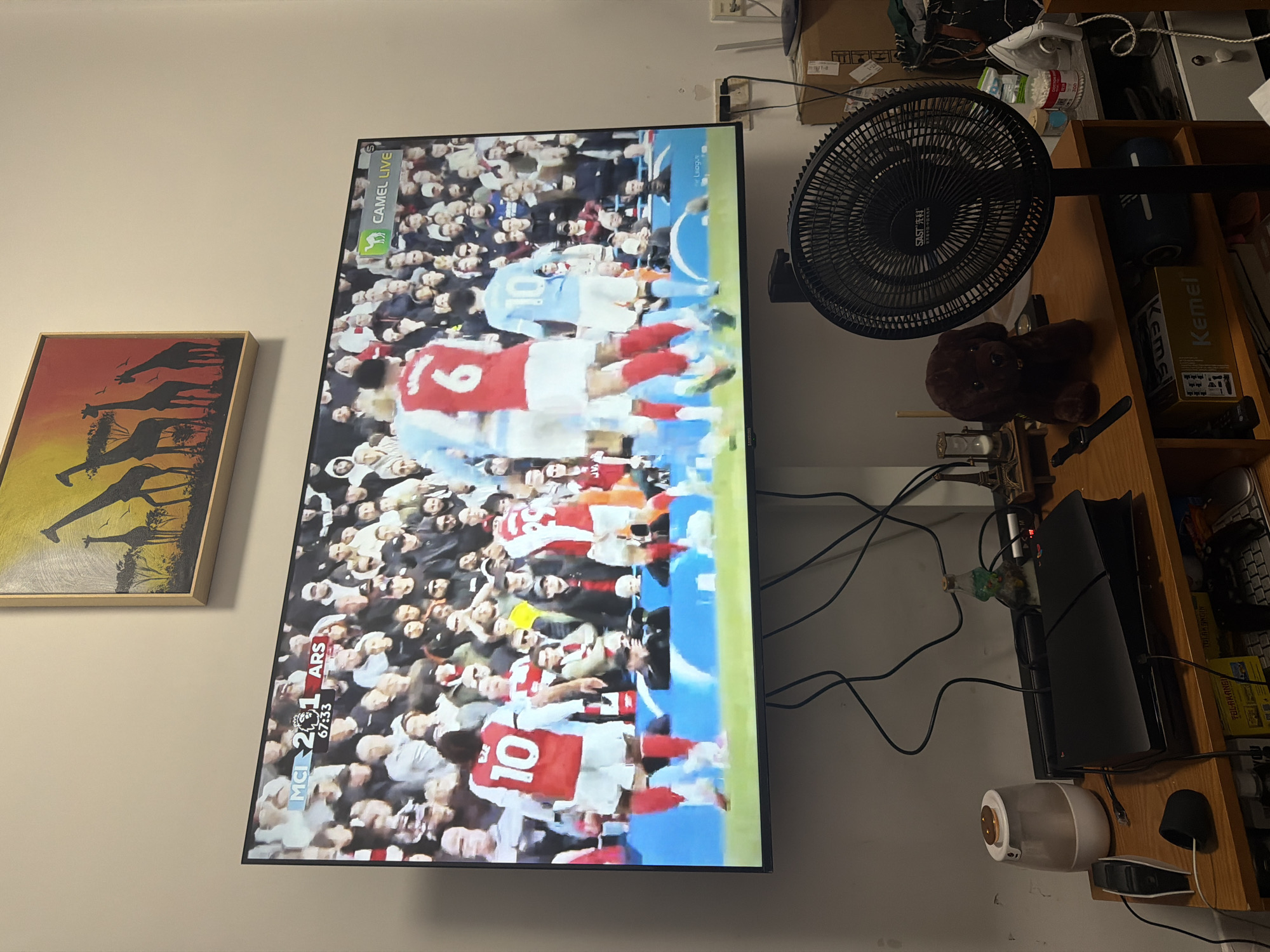 A television is on, displaying a soccer game between Manchester City (MCI) and Arsenal (ARS). The score is 2-1 in favor of Arsenal, with the game clock reading 67:33. Visible on the screen are players wearing the numbers 6 and 10. To the right of the TV is a black fan, shelves stacked with various items, including a Kemei box and a Sony PlayStation 5 console. Cords from the TV and other electronics are tangled. To the left of the TV is a framed painting or print featuring giraffes against a sunset. The word "SAST 先科" is visible on the fan. It is impossible to confirm the location in the city of Jiangning District, China from this image.