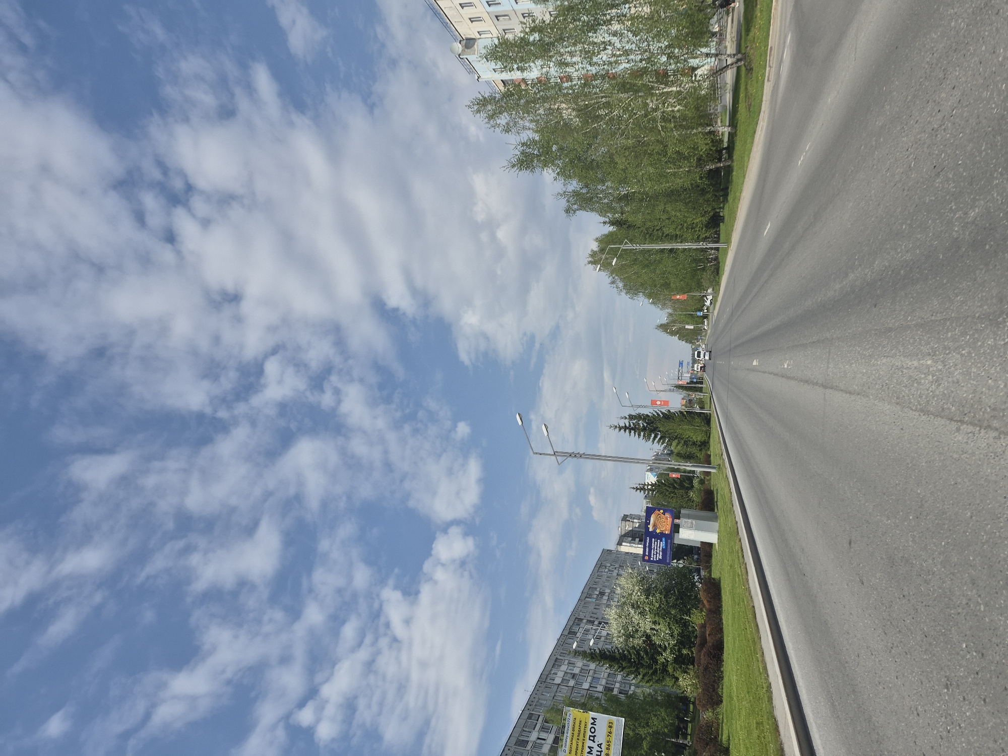 The image depicts a street in Tobolsk, Russia, under a blue sky with scattered white clouds. A gray asphalt road stretches into the distance, flanked by green grassy areas and trees. Several streetlights line the roadside. Buildings are visible along both sides of the road. A billboard is positioned near the road.