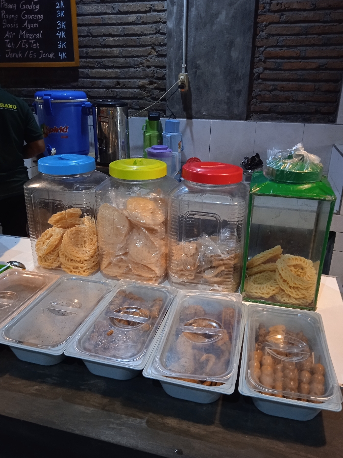 The image shows a food stall in Surakarta, Indonesia, located at coordinates -7.567 latitude and 110.799 longitude. The stall is situated in a brick-walled room. The focal point of the image is a row of five clear plastic containers filled with various foods. The first container holds a stack of fried pastry squares. The second container is filled with a similar type of pastry, but the squares are larger and more irregular in shape. The third container also holds a stack of fried pastry squares. The fourth container holds small, flat, round fried pastry. The fifth container holds what appears to be small, round, cooked snacks. The food items are arranged in a neat and organized manner. Behind the containers, there are three larger containers with lids, each filled with different types of fried snacks. These are arranged neatly on a counter. One of the containers has a blue lid with the word "Spirit!" printed on it. There is a partially visible figure on the left edge of the image. The figure is wearing a black shirt. The back wall of the stall is adorned with a blackboard displaying a price list in Indonesian. There is a wall-mounted switchboard on the wall. The scene is clean and well-lit. The lighting is artificial, coming from a source that is not visible in the image.