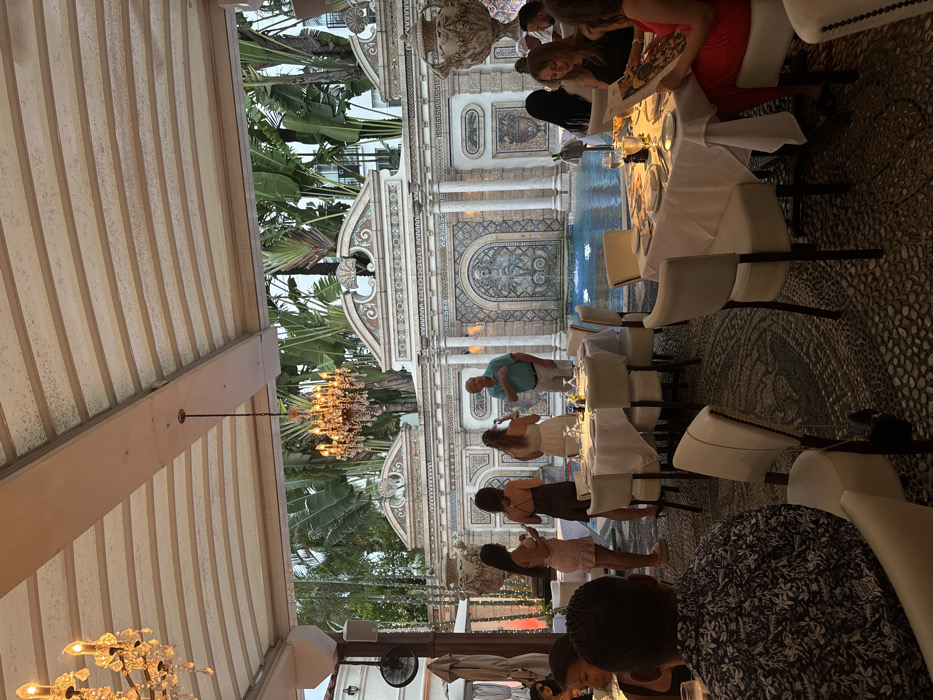 The image captures an outdoor dining scene at The Villa Casa Casuarina, located at 1116 Ocean Drive, Miami Beach, United States. The setting is an open-air covered patio beneath a striped fabric canopy from which two illuminated crystal chandeliers are suspended. A dark-colored ceiling fan is mounted on a structural beam to the left.
The floor is a dark-toned mosaic or cobblestone pattern. Several round dining tables, each covered with a white tablecloth, are arranged throughout the area. Each table is set with white plates, various glassware, and cutlery, with some featuring small condiment bottles. Guests are seated in white upholstered chairs with dark wooden frames.
In the background, an ornate, neoclassical-style wall made of light-colored stone and intricate mosaic tiling forms the perimeter of a blue-tiled swimming pool. A central fountain within the pool is active, emitting water jets, and features a mosaic design. Two large, decorative urns are positioned on top of the wall. Behind the wall, lush tropical vegetation, including large banana tree leaves, is prominent, with parts of a distant building visible through the foliage.
Approximately 15-20 individuals are present. In the mid-ground, a group of four stands near the pool's edge: a light-skinned male in a light blue collared shirt gestures with his right hand while facing three women. One woman wears a white strapless dress, one a dark sleeveless dress, and another a light-colored short-sleeved dress; each appears to be holding a mobile phone. Seated diners occupy the foreground and mid-ground. A male in the immediate foreground is visible from the back, wearing a dark patterned short-sleeve shirt. To the right, a woman with long dark hair, wearing a dark top, is observed looking at a menu, with another woman in a red top partially visible to her right, also examining a menu.