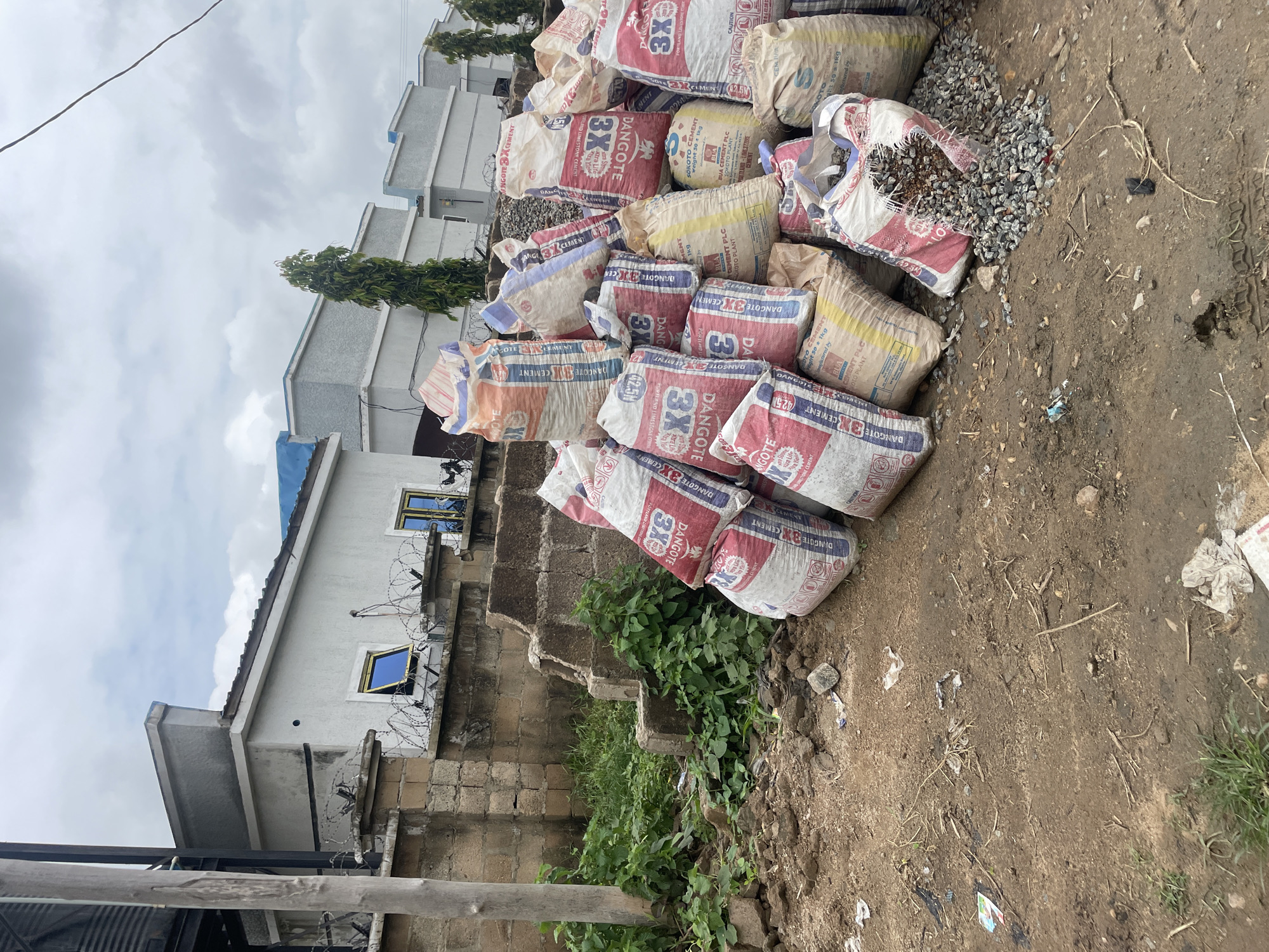 The image depicts a pile of cement bags and construction materials on a dirt lot in front of a row of modern, light-colored buildings with blue trim and solar panels on the roofs. The buildings have barred windows and are surrounded by a brick wall topped with barbed wire. The cement bags are mostly red and white, with various brand names visible, including "DANGOTE" and "SOROTO." Some bags are ripped, spilling out gravel and what appears to be aggregate. The ground is a mix of dry dirt, scattered debris, and patches of green grass. The sky is overcast with gray clouds. The scene appears to be an outdoor construction or storage area in a developing urban environment. The location is identified as Unknown, Nigeria.
