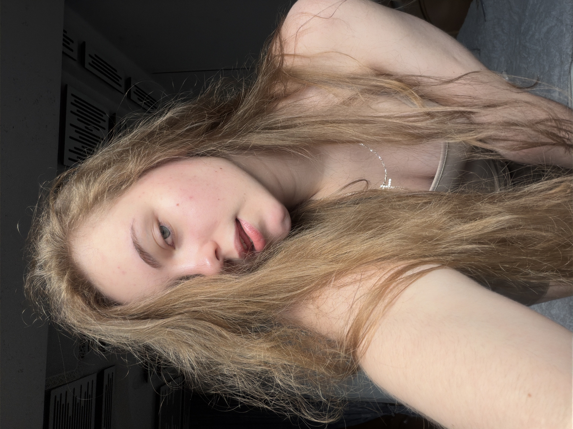 A young woman with long, blonde hair is shown in a close-up portrait. She is wearing a light-colored top and a silver necklace with a cross pendant. Her gaze is directed towards the camera, and her head is tilted. The background is dark and indistinct, with some horizontal lines visible on what appears to be a ceiling or wall fixture. There is no discernible location in the city of Dnipro, Ukraine, or any specific environmental context. The image is a selfie-style photograph.