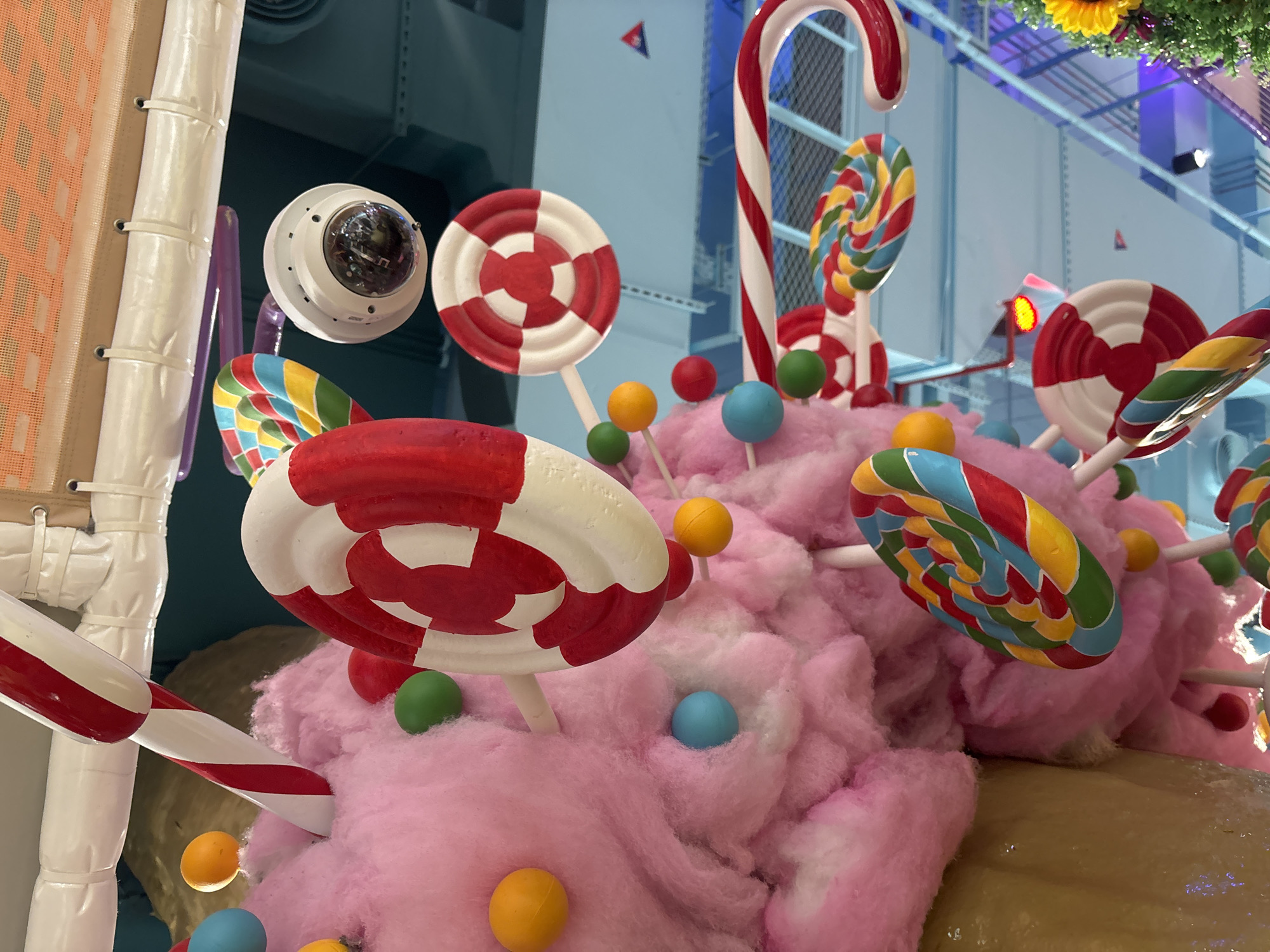 The image shows a whimsical, candy-themed decoration hanging from the ceiling. The decorations include large, swirling lollipops in various colors, candy canes, and what appears to be a giant swirl of pink cotton candy with small colorful balls embedded in it. A security camera is visible among the decorations. The background suggests an indoor environment with blue-toned structures, possibly a mall or entertainment venue. This scene is likely part of the decor for a seasonal event or a themed attraction in Dubai, United Arab Emirates, given the over-the-top and festive nature of the display. There are no people visible in the image.