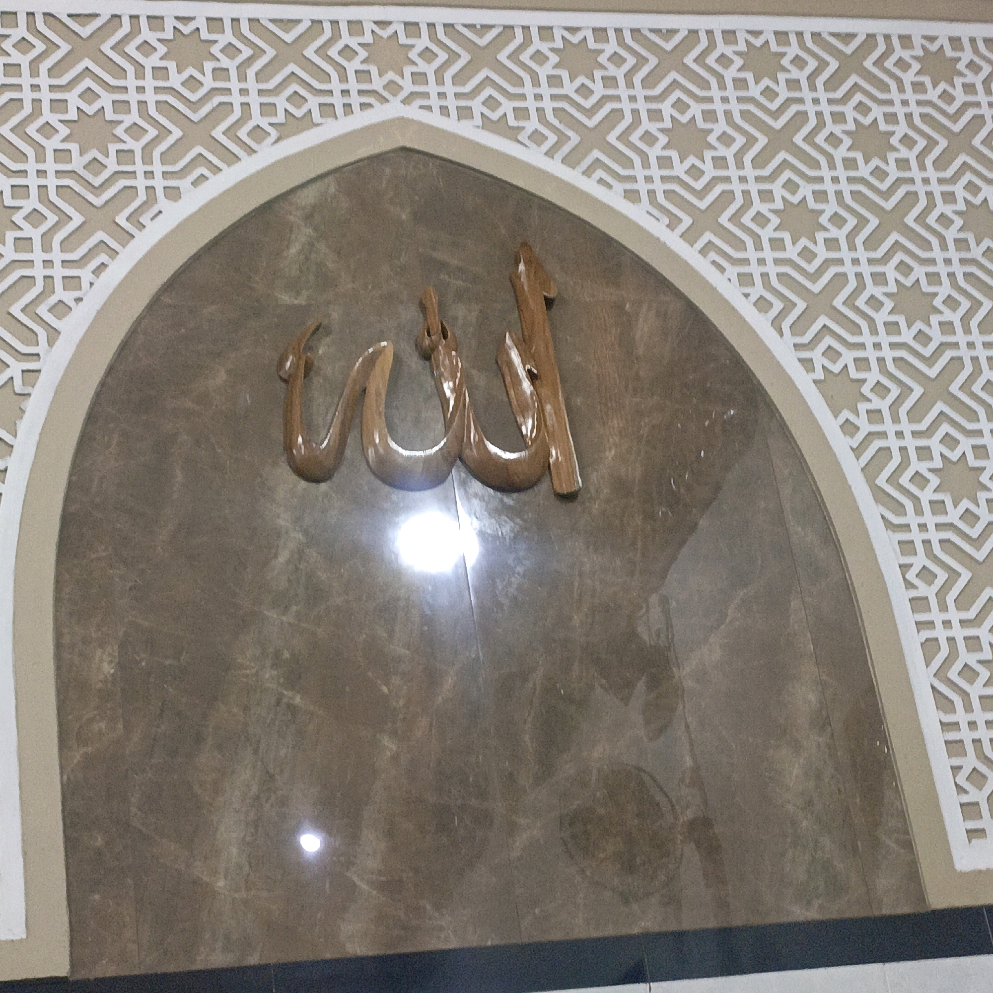 This image shows a close-up of an Islamic calligraphy that says "Allah" in Arabic script, carved from wood and mounted on a marble wall. The calligraphy is set within an arched alcove, which is surrounded by a decorative geometric pattern. The lighting in the image creates reflections on the marble surface. The exact location in Salatiga, Indonesia, is not identifiable from the image itself.