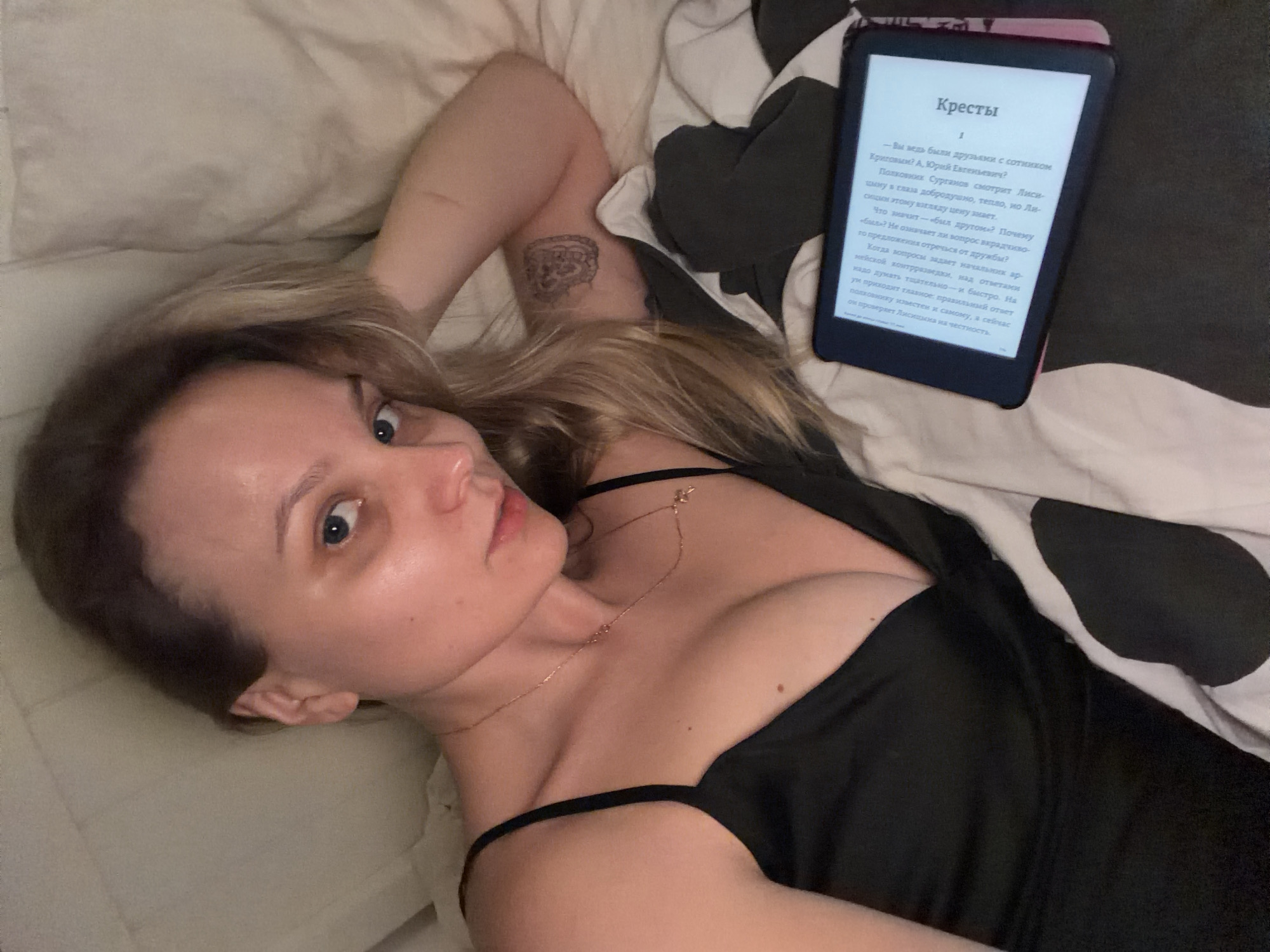 A woman is lying on a bed, looking at the camera. She is wearing a black top and a gold necklace. Her left arm is raised and visible, with a tattoo on the inside of her upper arm. An e-reader sits to her right, displaying a page of text in Russian. The bedding has a mix of white and patterned gray fabric.
There is no information available in the image to describe the location in Tel-Aviv, Israel.
