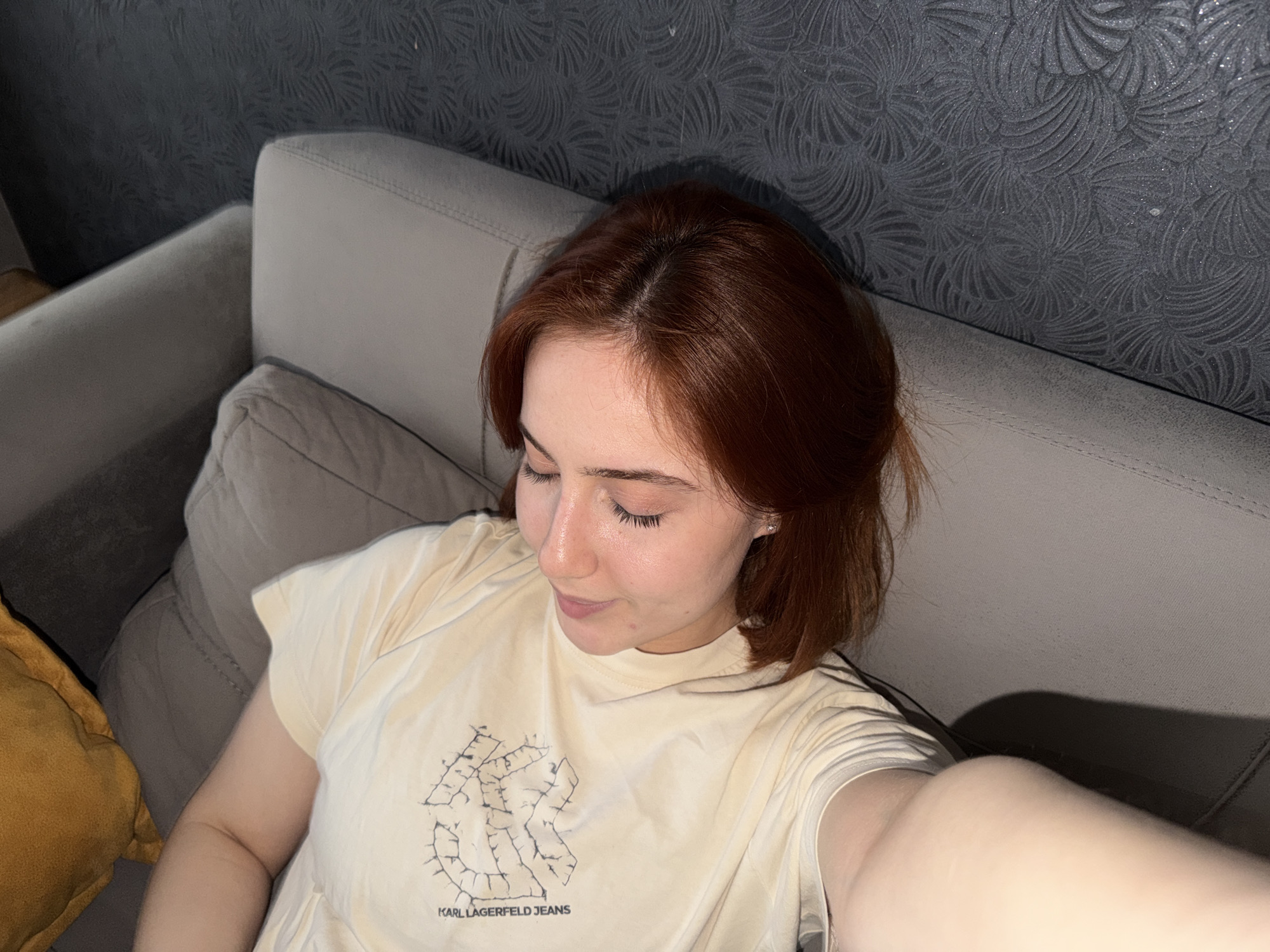 A young woman with red hair is reclining on a grey sofa. She is wearing a light yellow t-shirt with a black logo on the front. The background is a grey wall with a textured pattern. The photo appears to be taken from a high angle, possibly as a selfie. A yellow pillow is visible in the lower left corner of the image. The location is not discernible as Tbilisi, Georgia from the visible content.