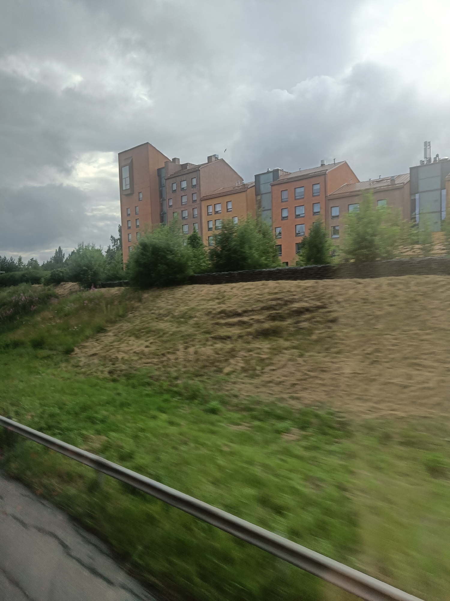 This media file captures a view of a modern, multi-story building complex on a cloudy day. The building features orange-toned facades and numerous rectangular windows, with some sections having a contemporary glass and metal design. Trees and foliage are visible in front of and around the building, suggesting a suburban or park-like setting. A gently sloping grassy embankment runs across the foreground, with a metal railing visible along the bottom edge of the frame, indicating the perspective might be from a moving vehicle or a walkway. The sky is overcast with dramatic clouds, and a faint ray of sunlight breaks through on the right side, suggesting it's either early morning or late afternoon. The overall impression is one of an urban landscape with natural elements, under a somewhat somber sky. There is no visible text in the image.