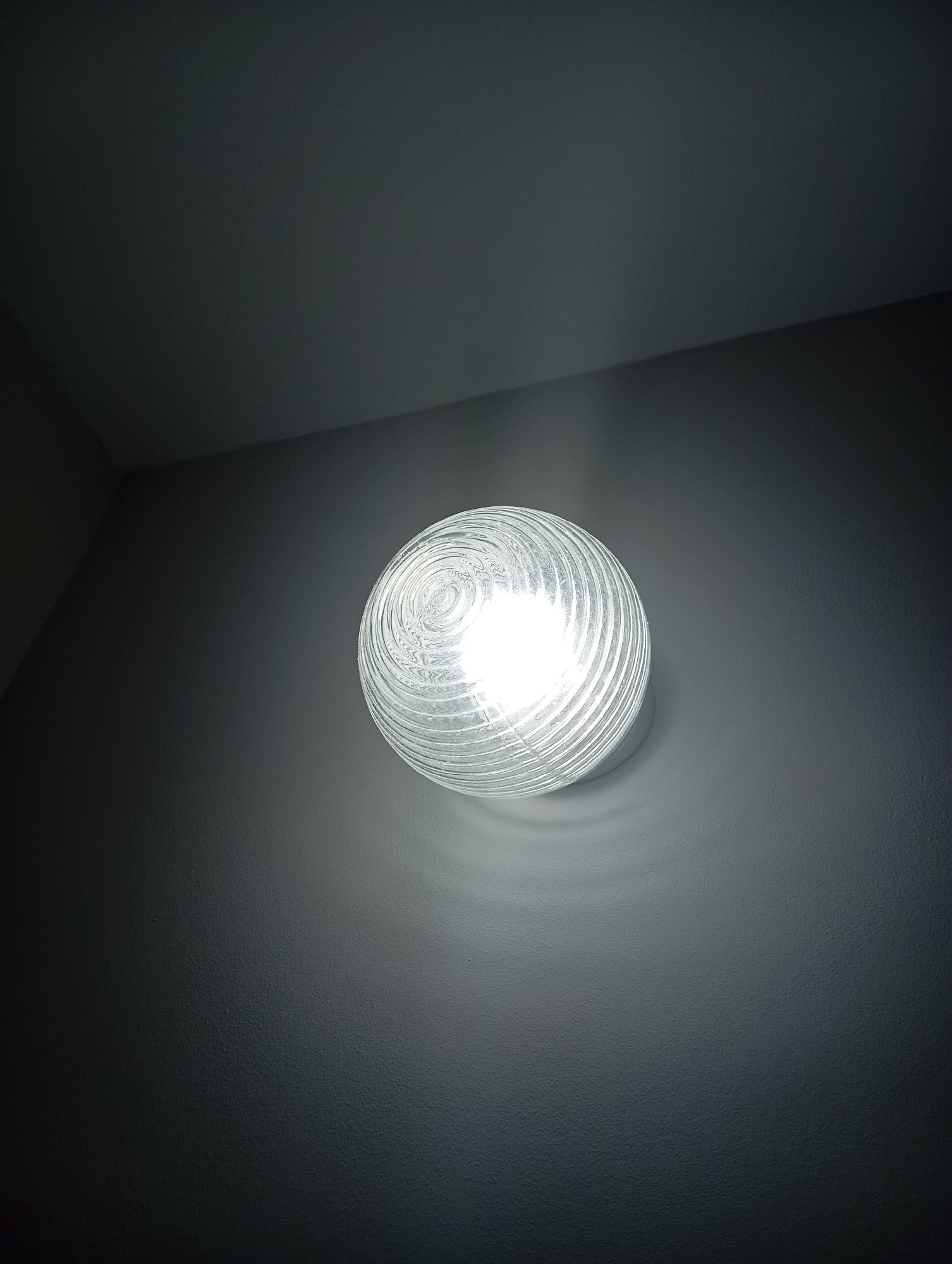 An indoor, low-angle shot shows a single, lit, clear glass light fixture on a dark gray wall. The light fixture is spherical and has a textured surface with concentric, raised rings. The bulb inside is bright white and illuminates the fixture from within, casting a soft glow on the surrounding wall. The wall appears to be painted or textured and is darker in the upper left and lower right of the frame due to the lighting. The ceiling is visible in the upper portion of the image and is also dark gray. There are no people or other objects in the scene.