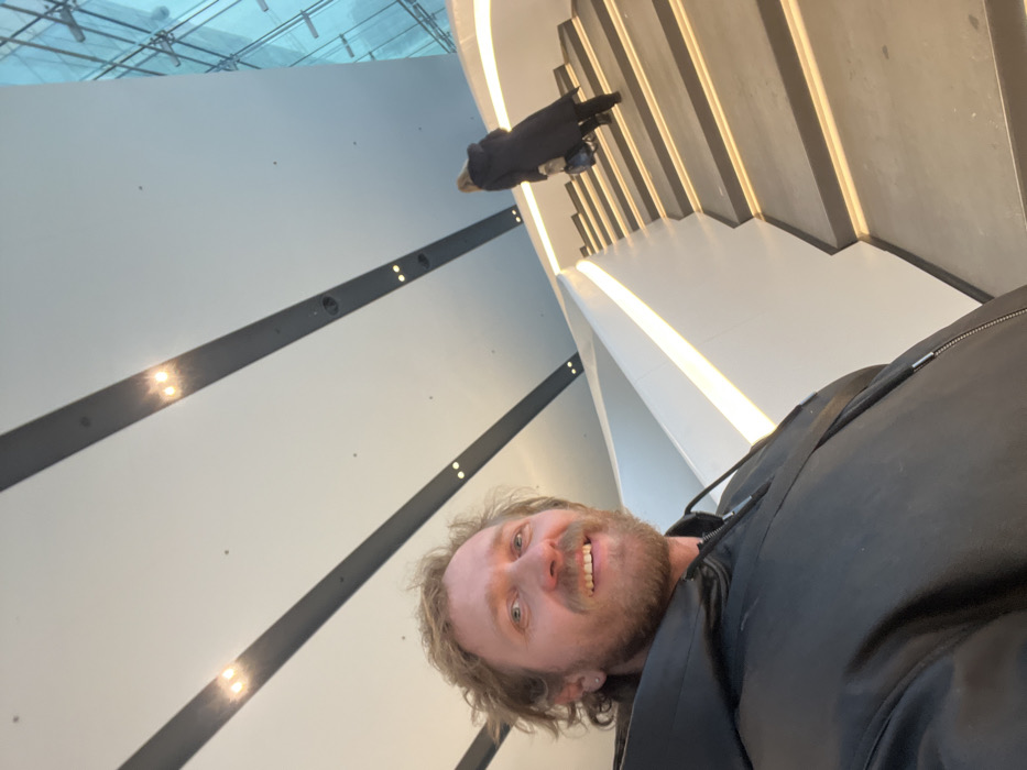 The image displays an interior scene within a modern building, identified as the Lakhta Center in Saint Petersburg, Russia.
In the foreground, a male individual with light brown, curly hair and a beard smiles directly at the camera, suggesting a selfie. He wears a black hooded jacket with a visible zipper and a dark strap across his chest. A small earring is visible in his left ear.
Behind the male and to the right, a female individual is observed ascending a wide, contemporary staircase. She is seen from the back, has light-colored hair, and wears a long, dark coat and dark boots, carrying a dark bag in her left hand.
The staircase features a minimalist design with solid white balustrades and steps. Each step is illuminated by a horizontal strip of warm white LED lighting integrated along its front edge, and a similar strip runs along the upper edge of the balustrade. The ceiling above is high and light-colored, traversed by parallel dark structural beams or decorative strips, each containing multiple recessed circular spotlights. To the far right of the frame, a large, multi-paneled glass wall with a distinct blue tint and visible internal framing suggests an expansive exterior view or atrium.