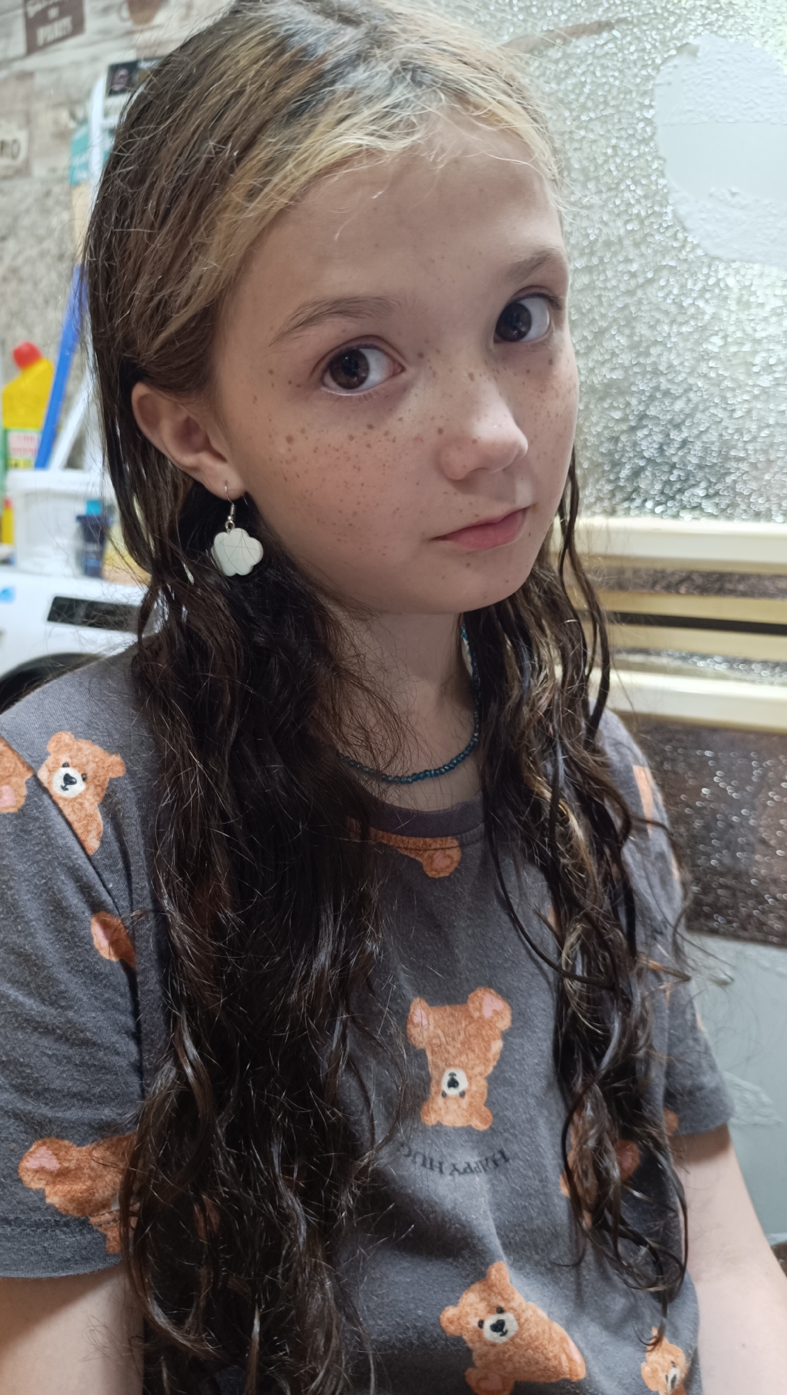 A young girl with long, dark, curly hair and freckles is pictured in a close-up shot. She has brown eyes and is looking directly at the camera with a neutral expression. She is wearing a gray t-shirt with a repeating pattern of brown teddy bears. She also has a beaded necklace and a white cloud-shaped earring in her left ear. The background appears to be indoors, with a textured, possibly frosted, window or glass partition to her right. Some objects are visible in the background on the left, including what looks like a bottle of cleaning product. The lighting suggests it might be daytime.