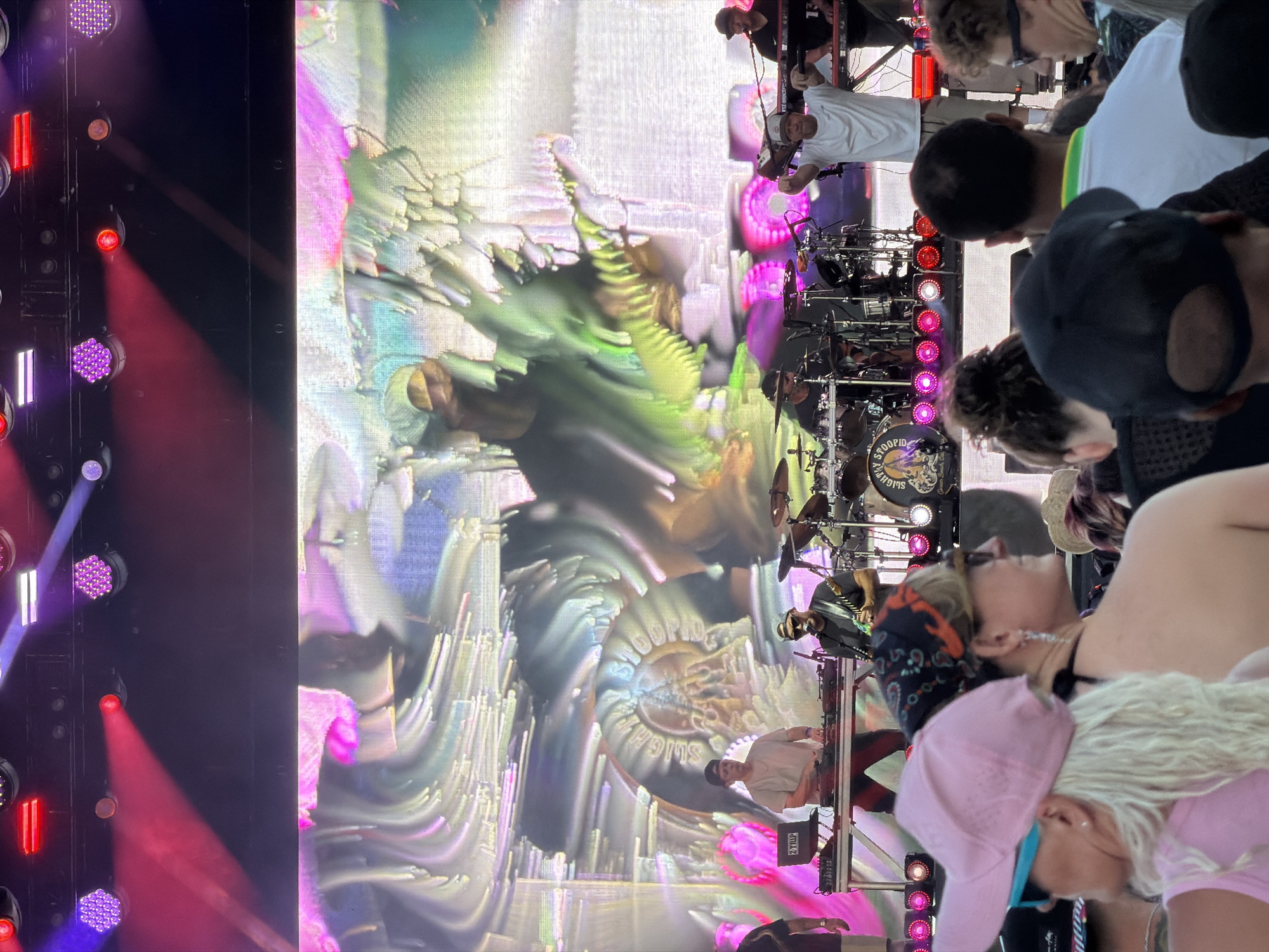 This is a medium shot, taken from behind a crowd of people, of a musical performance at an outdoor venue. The stage is lit with pink and red lights, and a large, colorful, abstract digital backdrop is displayed behind the performers. In the foreground, the backs of several audience members are visible, with one woman in a pink hat and blonde hair in focus on the left. On stage, from left to right, a person is standing behind a DJ setup. To their right, a person wearing a hat is playing a guitar. Further to the right, a drummer is seated behind a drum set. Another person stands behind a keyboard, and to their right, another person is also behind a keyboard. The overall scene suggests a lively concert atmosphere.