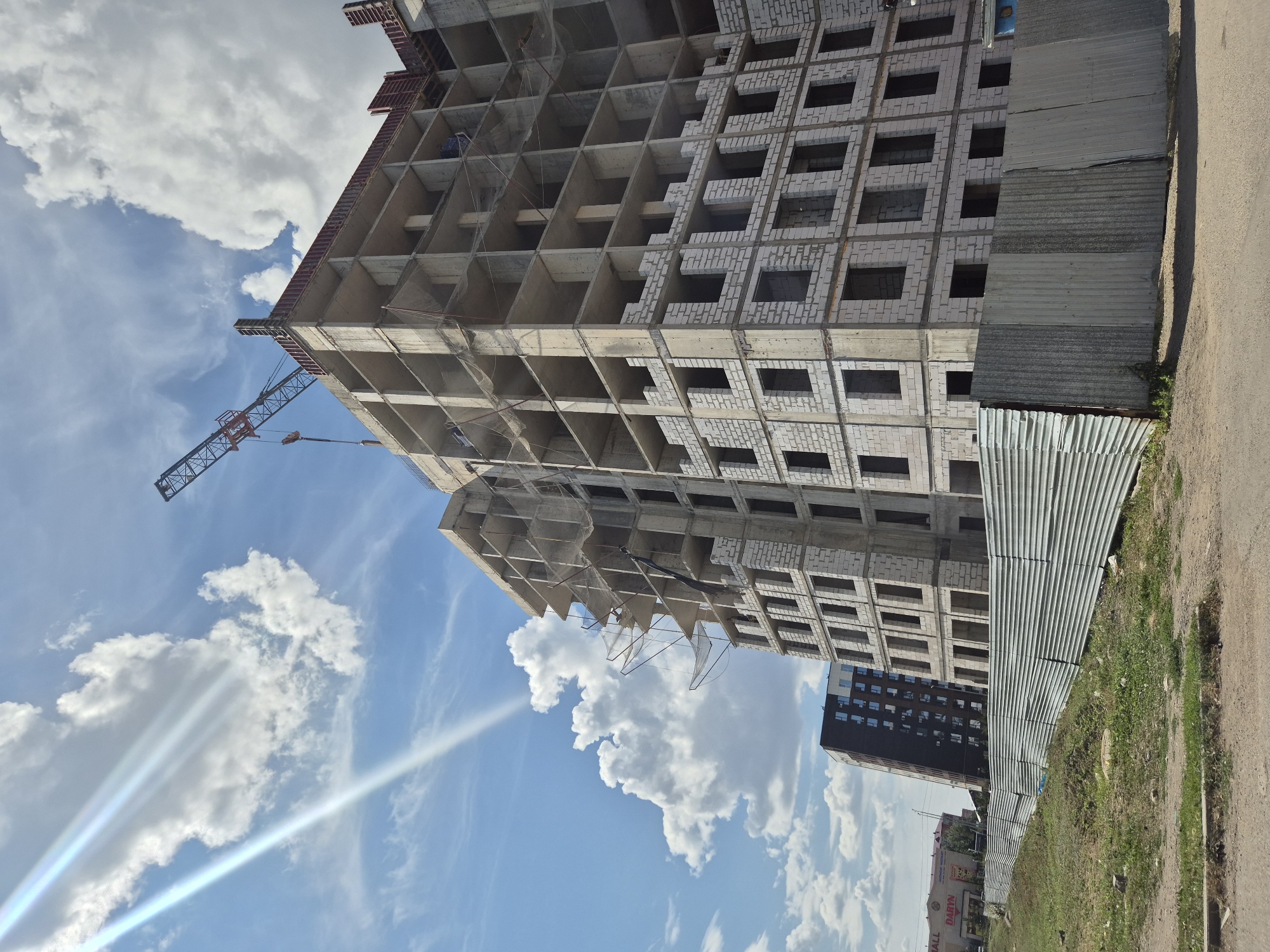The image depicts a construction site in Kokshetau, Kazakhstan, on a sunny day with scattered clouds. In the foreground, a corrugated metal fence runs horizontally across the scene, with a grassy embankment behind it. The main subject is a multi-story building under construction, characterized by its concrete frame and white brick infill. A construction crane is visible at the top of the building, with its arm extended. To the left of the main building, a dark, modern building with numerous windows is also under construction, leaning slightly. In the far left background, a shopping center with the word "DARYN" visible on its facade can be seen. The sky is blue with bright sunlight casting rays through the clouds. No people are visible in the image.
