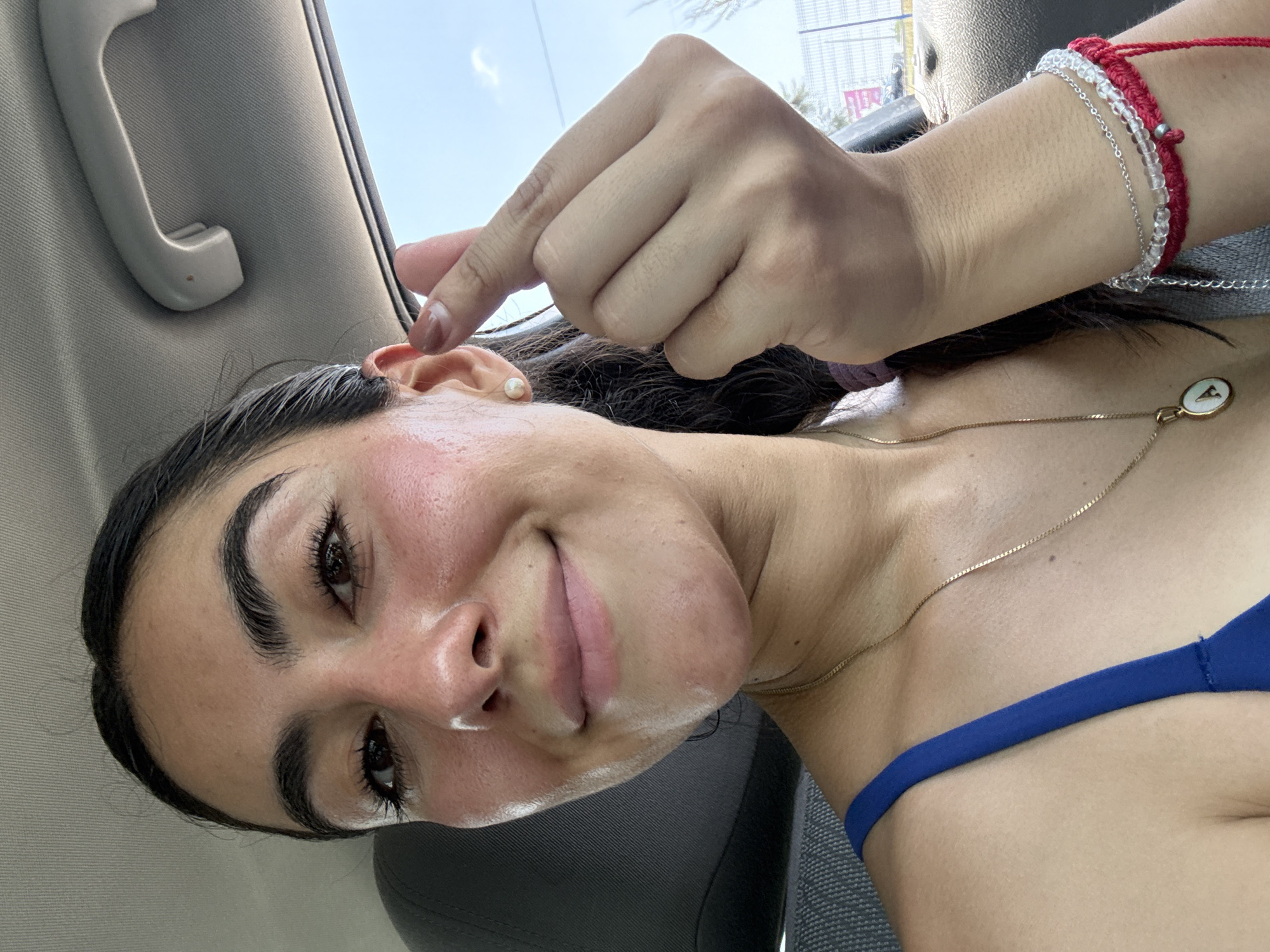 A young woman is inside a car. She is holding her left ear with her hand. She has dark hair, dark eyebrows, and long eyelashes. She is wearing a pearl earring and a gold necklace with a white circular pendant. She has a red string bracelet and a silver bracelet with clear beads on her left wrist. She is wearing a blue top. The interior of the car is gray. In the background, there is a building, a palm tree and the sky. The location is possibly the city of Puebla, Mexico based on the urban architecture and the presence of palm trees, which are common in the region.
