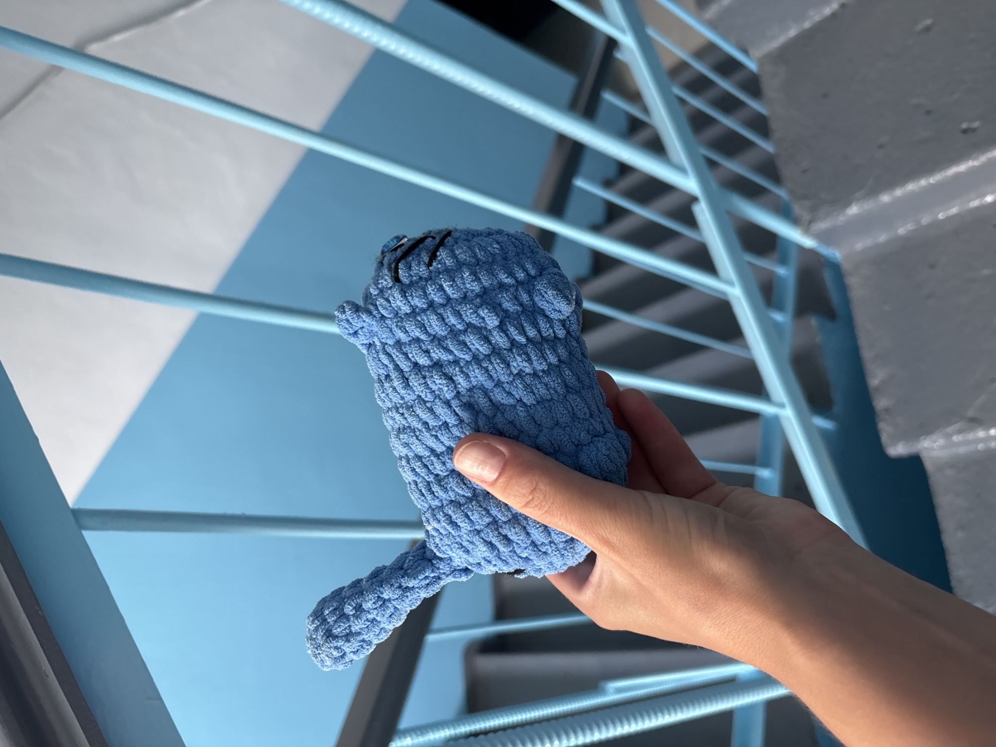 A person's hand is holding a small, light blue, crocheted toy animal, possibly a cat, in front of a set of light blue metal stairs. The toy has black embroidered eyes and eyelashes. The stairs are part of an outdoor staircase, with grey concrete steps visible below. The background consists of light blue and white panels, creating a diagonal pattern. The overall scene suggests an urban environment, potentially a public space in Blagoveshchensk, Russia.