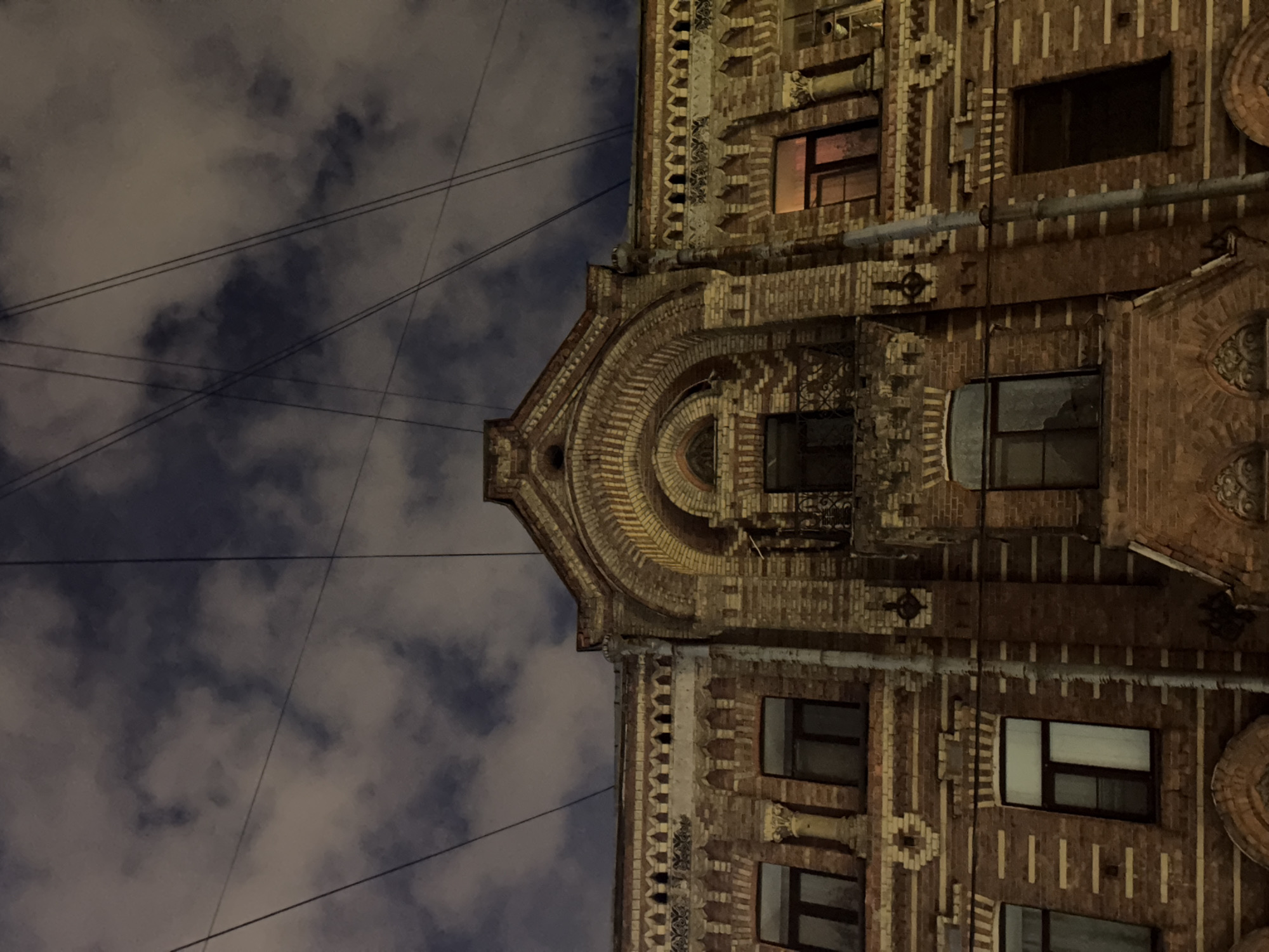 An upward-angle exterior shot captures the facade of an ornate, red brick building at night in Saint Petersburg, Russia. The building features multiple stories with numerous windows, some illuminated by an orange glow from within. Decorative brickwork and architectural details, including arched windows and cornices, are visible. Overhead, a dark, cloudy sky is crisscrossed by a web of utility wires.