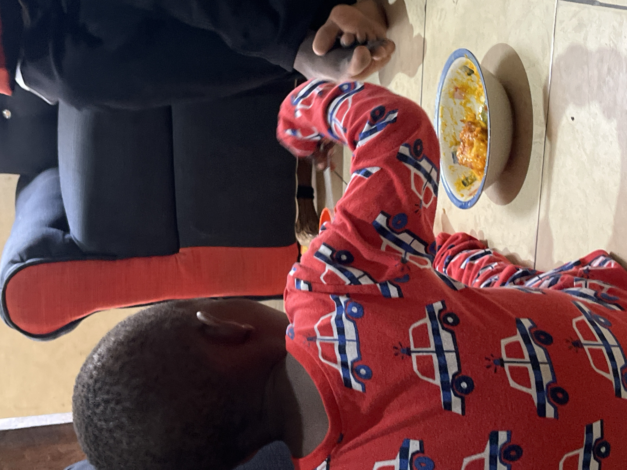A child wearing red pajamas patterned with white and blue cars lies on a tiled floor next to an adult. The adult is sitting on a chair with red trim and wearing dark clothing, with hands clasped. Next to the child is a dish with remnants of food. The location is possibly a home in Badagry, Nigeria, given the request.
