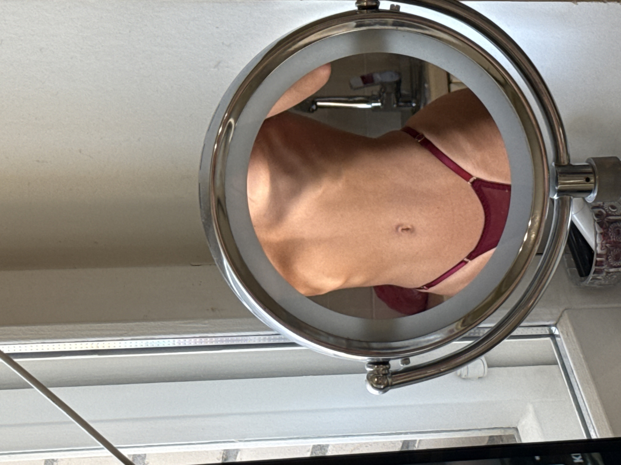 This is a close-up, medium shot of a person's torso, reflected in a circular makeup mirror. The person is wearing a burgundy thong. The mirror is framed by a silver metal stand, and is positioned on a surface that appears to be a vanity or dresser. The background includes a window and a wall. The image is well-lit, with the light source appearing to be from the window. There is no specific indication of the location being Saint Petersburg, Russia.