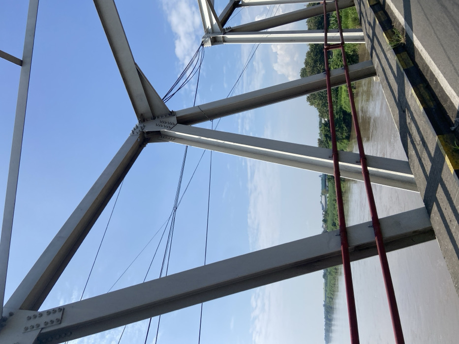 The image shows a close-up view of the underside of a white metal bridge. The bridge is located at the coordinates -7.486, 112.561 in Pungging, Indonesia. The bridge is over a brown river. There is a red railing on the side of the bridge. There is a clear blue sky with white clouds and several electrical wires running across the frame. There are trees on the banks of the river. The bridge is made of white metal and the structure is made of several large triangular beams. The image is taken from a perspective directly below the bridge. There are no people visible in the image.