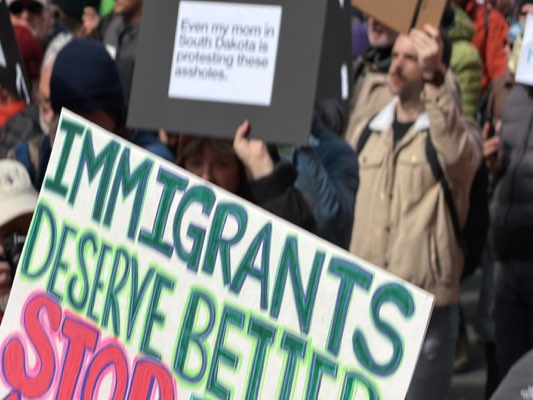 The image depicts a protest or demonstration. In the foreground, a sign reads "IMMIGRANTS DESERVE BETTER STOP" with "IMMIGRANTS DESERVE BETTER" in green letters and "STOP" in red. Behind the sign, people are holding another sign that reads "Even my mom in South Dakota is protesting these assholes." A man in a tan jacket stands behind the sign holders. The image is somewhat blurry. The location is not explicitly identified in the image.