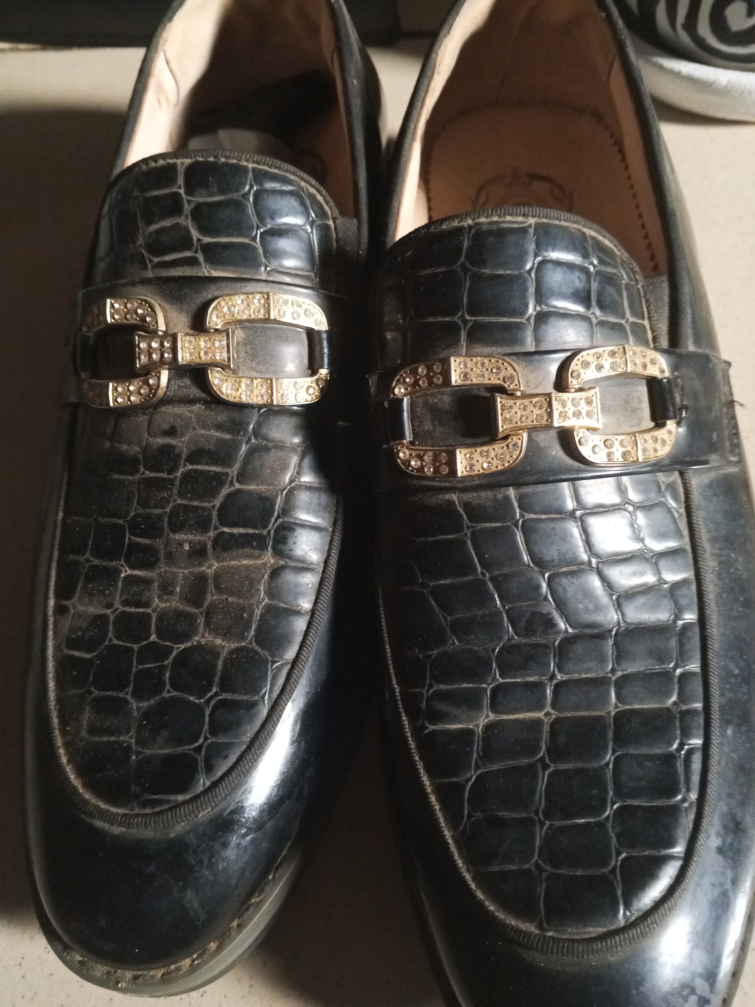 The image shows a close-up, overhead view of a pair of black, alligator-textured loafers with gold and diamond-encrusted embellishments. The shoes are dusty, indicating they may not be new or have been stored for some time. One shoe is slightly to the left and the other is slightly to the right, both positioned side-by-side on a neutral surface. The background is dark and indistinct, offering no specific environmental clues. There are no people visible in the image, nor any indication of the location being within Ashata, Nigeria.
