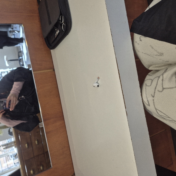 An indoor scene depicts a person's point of view from a chair in what appears to be a hair salon or similar grooming establishment. In the lower foreground, the individual's knees and thighs are visible, clad in light-colored trousers with a dark, abstract pattern, partially covered by dark fabric. A white, flat counter surface occupies the midground. Centrally placed on this counter is a single white Apple AirPod Pro earbud. To the right side of the counter, a black zippered case containing various hairdressing tools, including scissors, is visible. Above the counter, a large mirror with a light-brown wooden frame reflects the upper body of the person taking the photograph; this individual is wearing a dark protective cape over light pink long-sleeved attire and holding a smartphone. The mirror's reflection also shows a multi-drawer wooden dresser or cabinet with brass handles, and blurred elements in the background consistent with a salon setting, such as seating or equipment, and the indistinct form of another person's legs. The precise location within the city of Strasbourg, France, cannot be determined from the visual information provided.