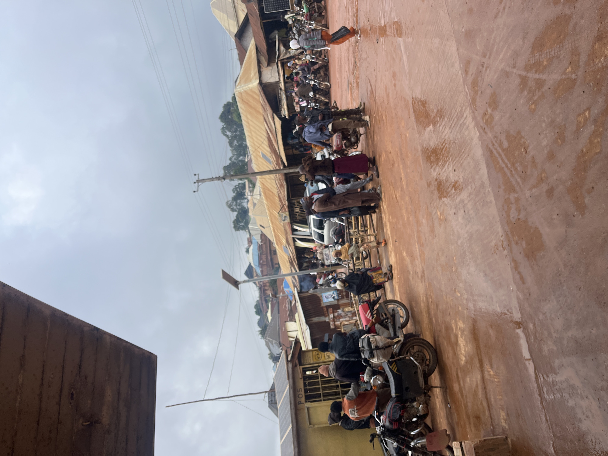 This is a sideways, slightly blurry, medium-shot photograph taken outdoors in what appears to be a marketplace or busy street in Gembu, Nigeria. The scene is dominated by a muddy, reddish-brown ground, suggesting recent rain or a dusty environment. A line of motorcycles, some with riders or passengers, are parked or moving along the street.
Several people are visible, some standing, some on motorcycles. They are dressed in casual clothing, including shirts, pants, and head coverings. Their expressions are not clearly discernible due to the image quality and distance, but the general atmosphere seems busy and perhaps a bit congested due to the parked vehicles and pedestrian activity.
In the background, there are various buildings with corrugated metal roofs, characteristic of many market structures in such regions. There are also utility poles and wires, indicating the presence of infrastructure. The sky is overcast, suggesting a cloudy or possibly rainy day.
One building has visible text that reads "POST" and another has a logo that appears to be "MTN". These offer clues about the types of businesses or services available in the area, such as postal services and a telecommunications provider.
The overall impression is of a bustling daily scene in a Nigerian town, with a focus on local transportation and commerce. The time of day is likely daytime, given the ambient light, but the overcast conditions make it difficult to pinpoint further.